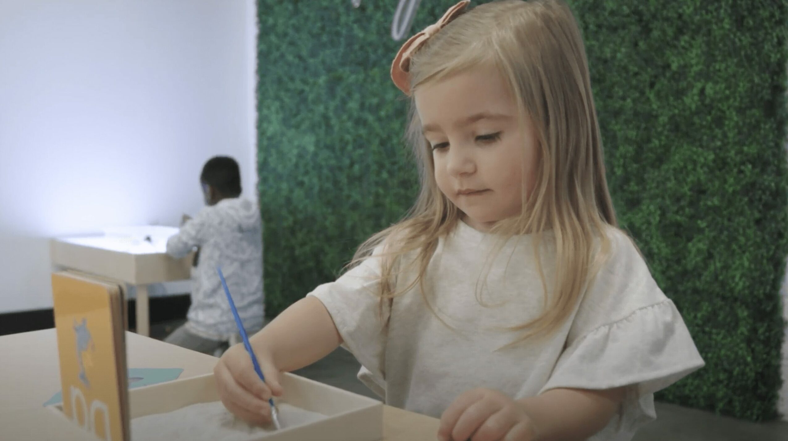 Young girl with long blonde hair writing with a blue pen at a desk, background includes a green wall and another child.