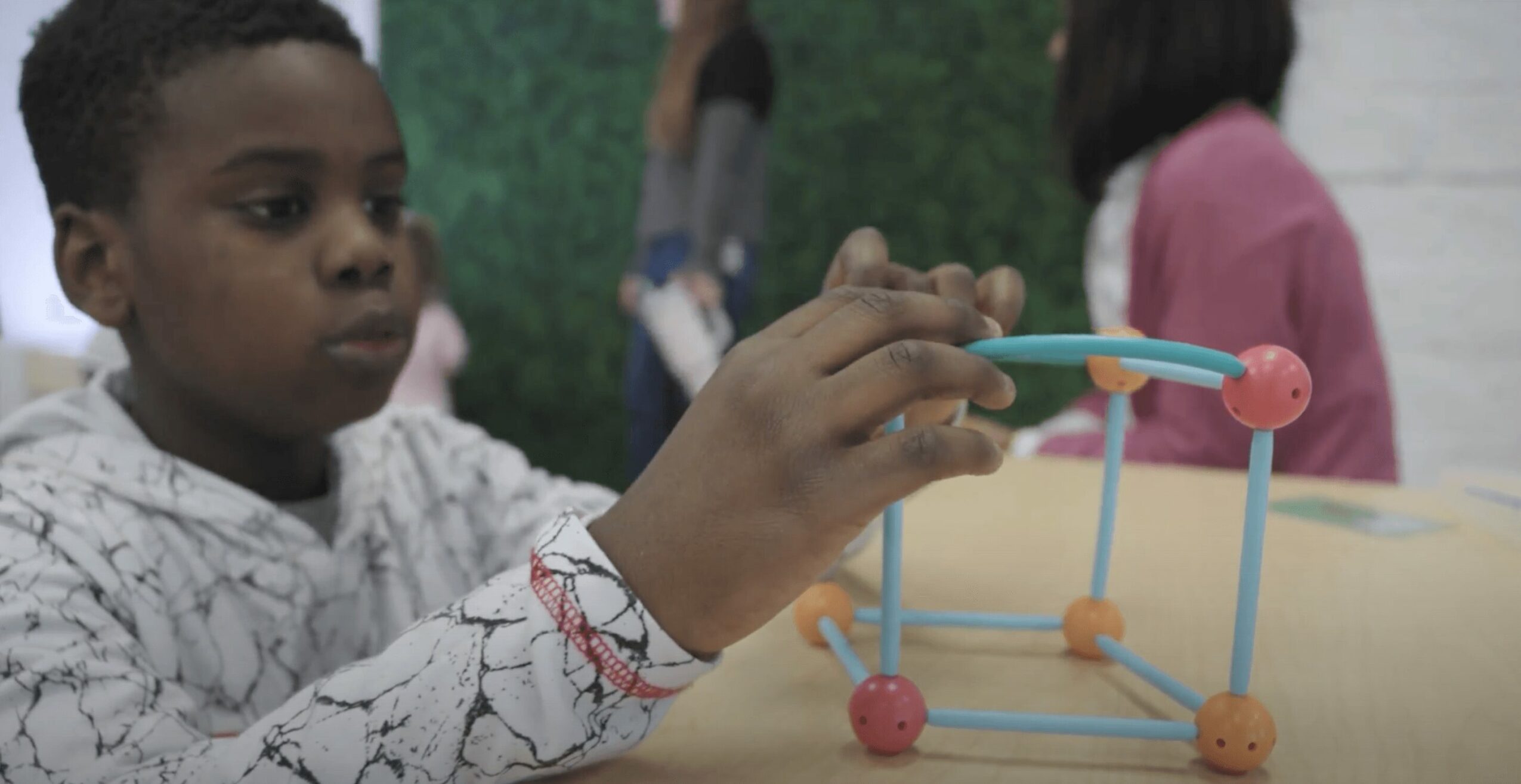 Child building with colorful geometric blocks at a table, with other children in the background.