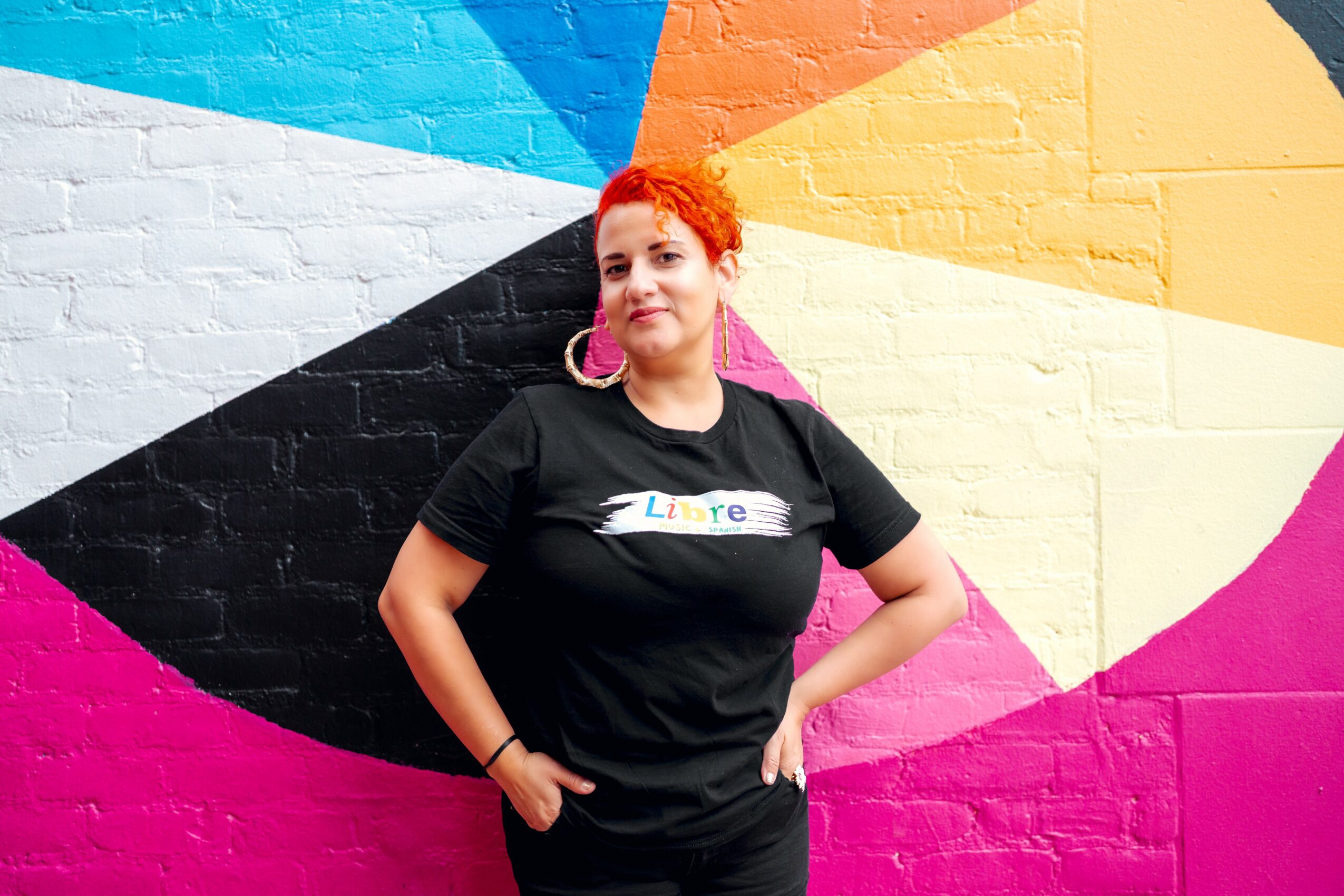 Woman with red hair and pink earrings stands in front of a colorful, geometric mural wall.