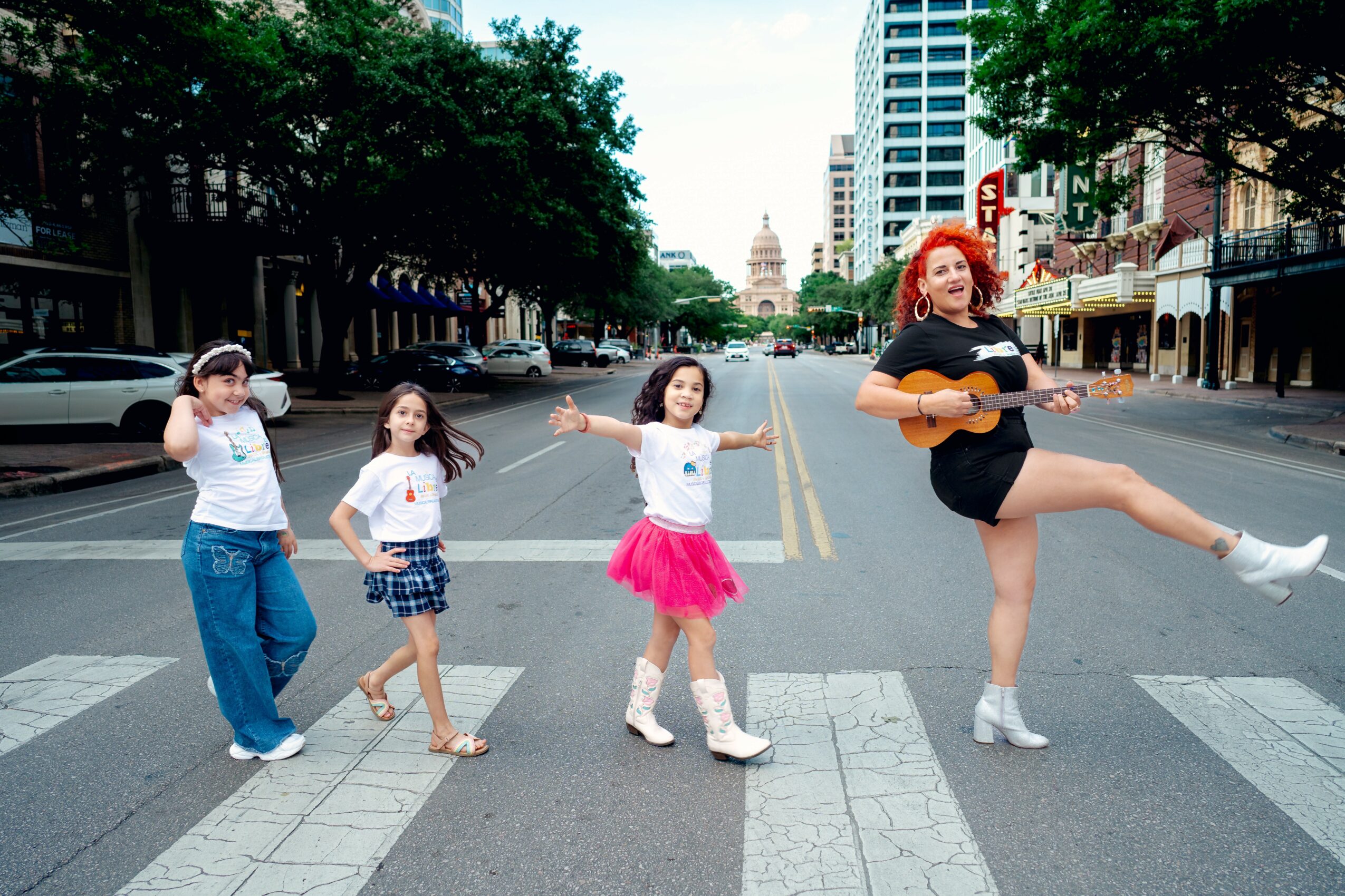 Four children and a woman with a guitar standing on a city street crosswalk, with buildings and trees in background.