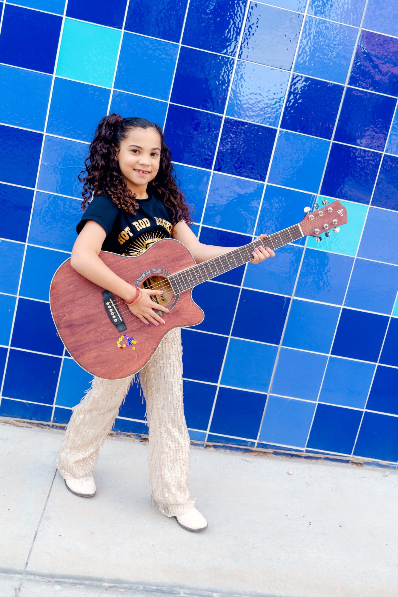 Girl with curly hair holding a guitar in front of a blue tiled wall.