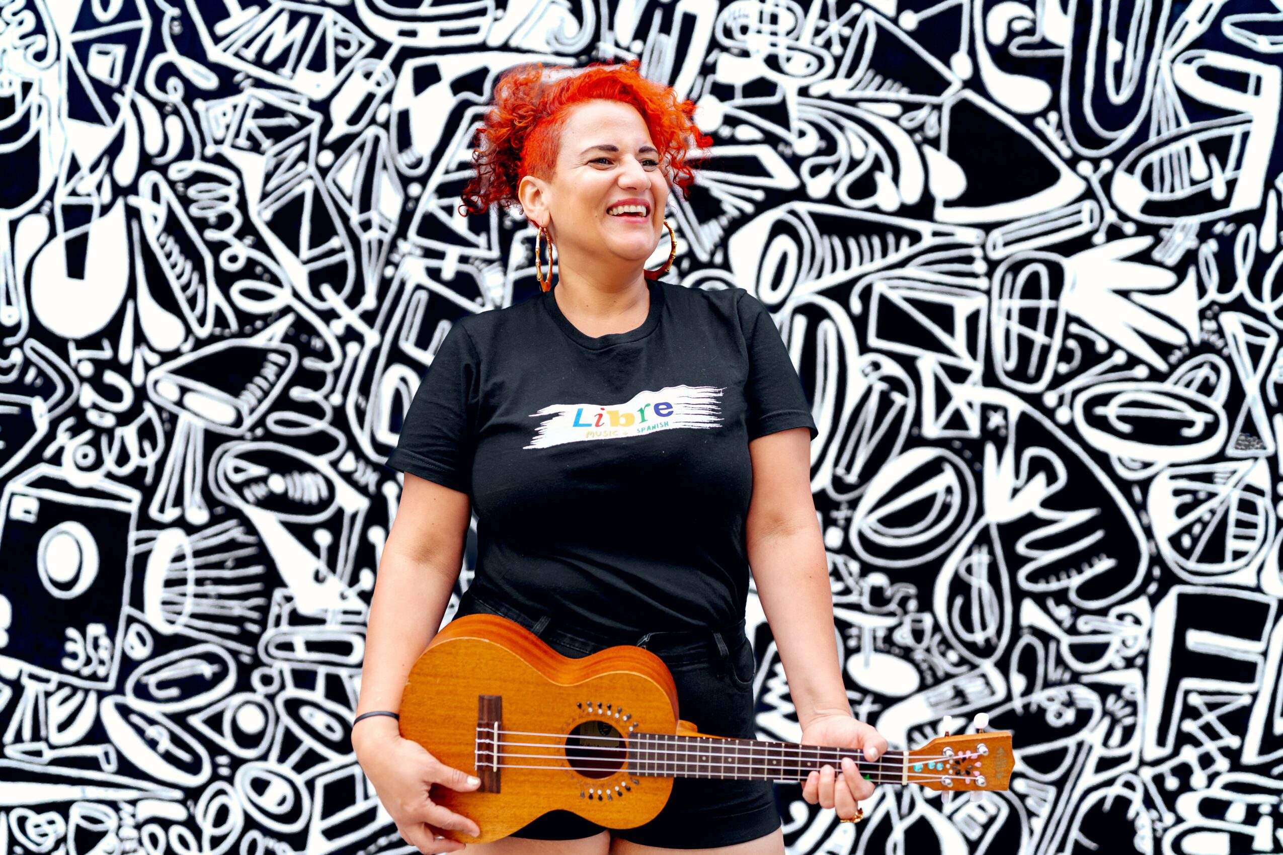 Woman with curly red hair holding a guitar, smiling, standing in front of a black and white abstract background.