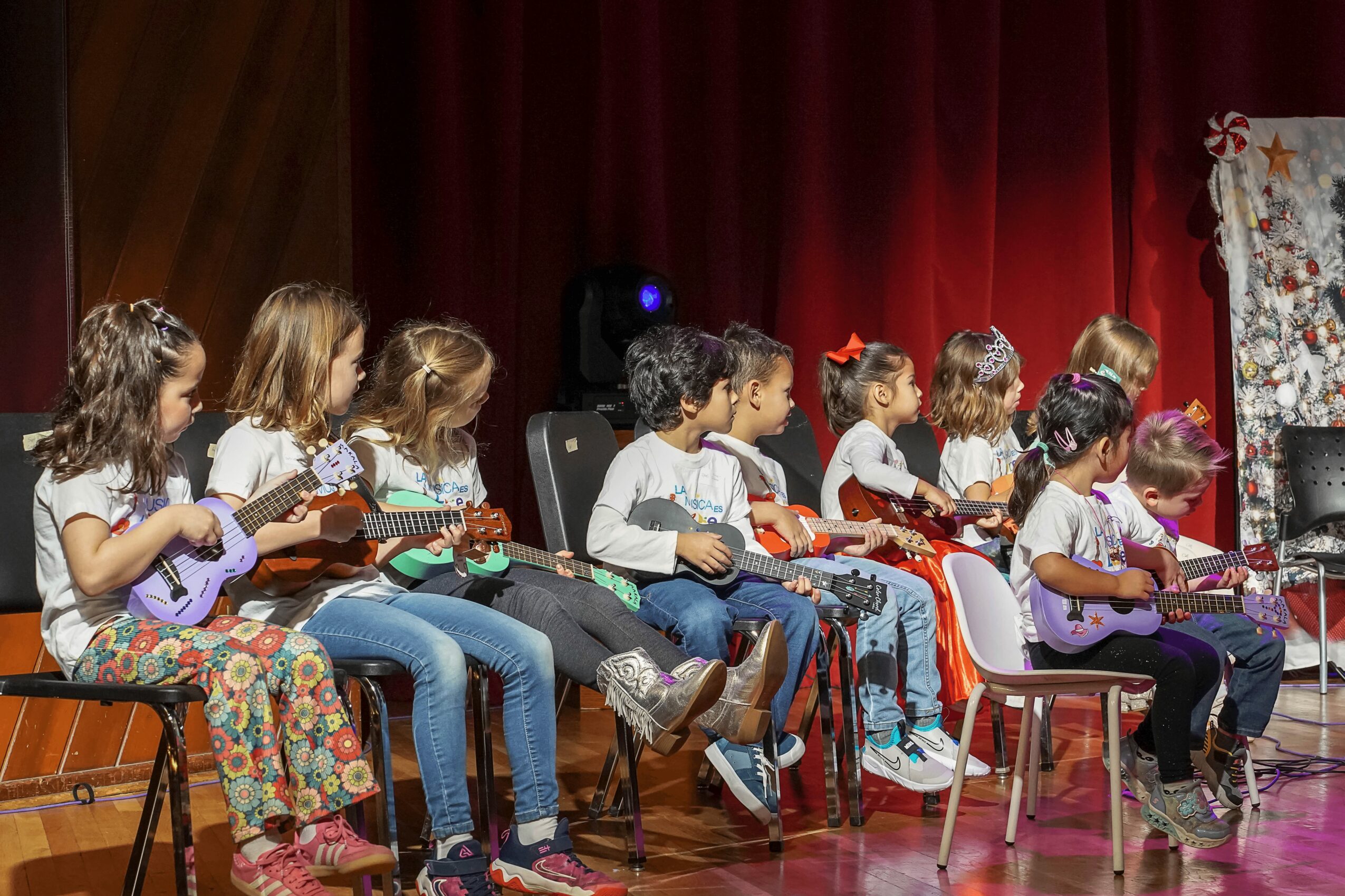 Children sitting on chairs playing ukuleles on stage with red curtain background.