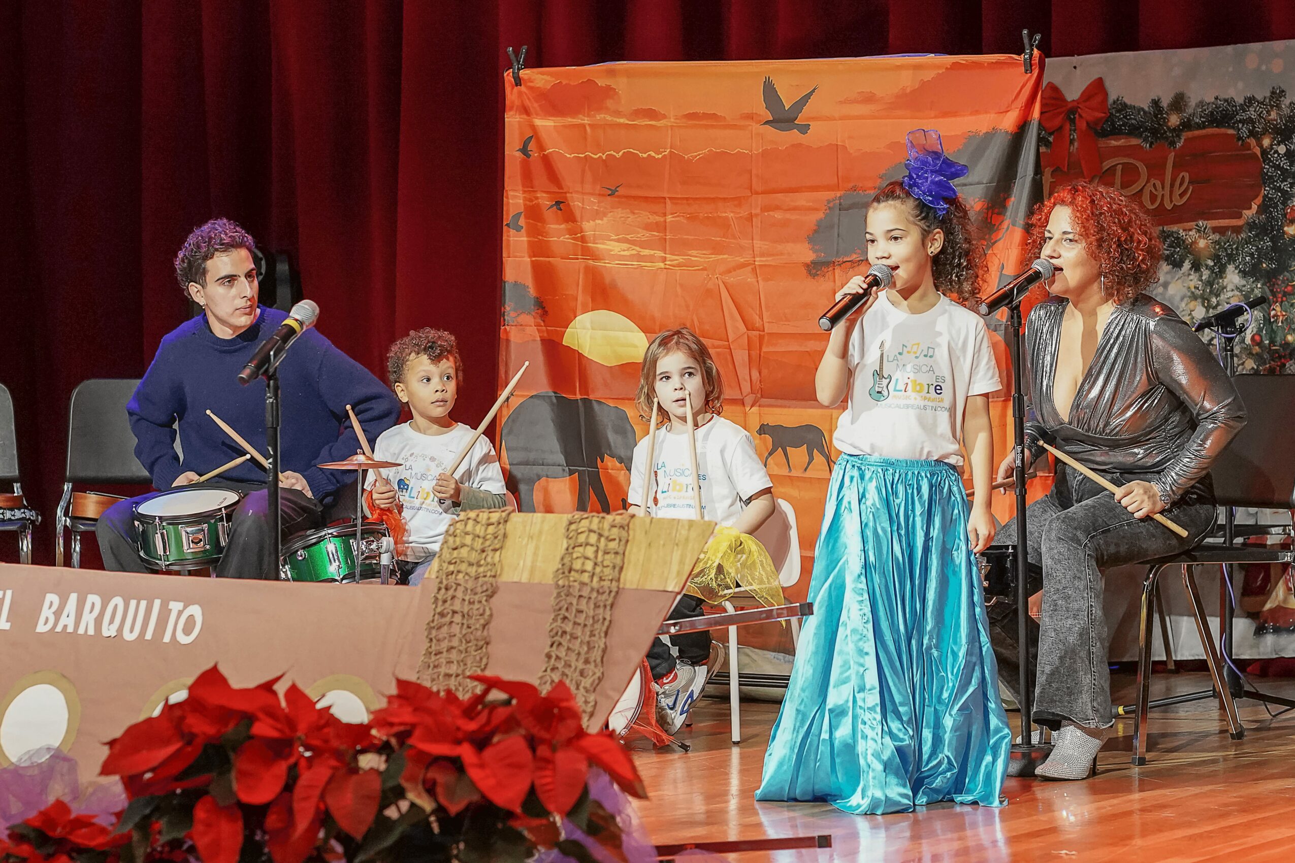 Group of children and two women performing on stage with red curtains and a colorful backdrop.