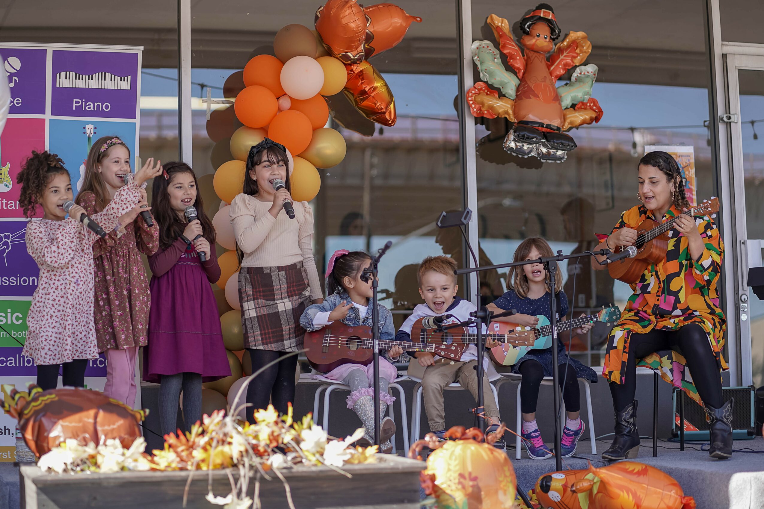 Children and a woman perform on stage with balloons and decorations, some children playing guitars, in a celebration setting.
