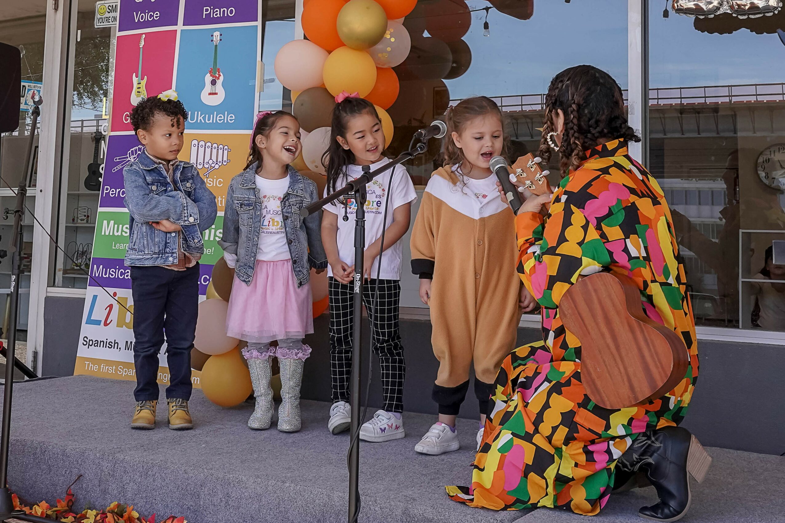 Group of five children standing on stage with a woman, balloons, and a microphone in front of a glass window.