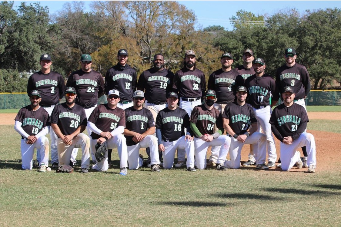 Group of baseball players in black and green uniforms posing on a field with trees in background.