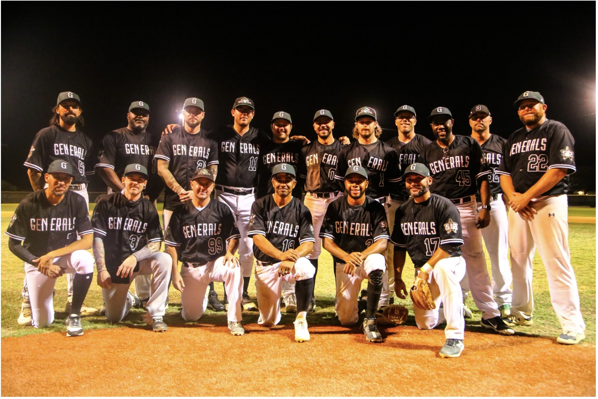 Group of baseball players in black uniforms with white pants, standing on a field at night, posing for a team photo.
