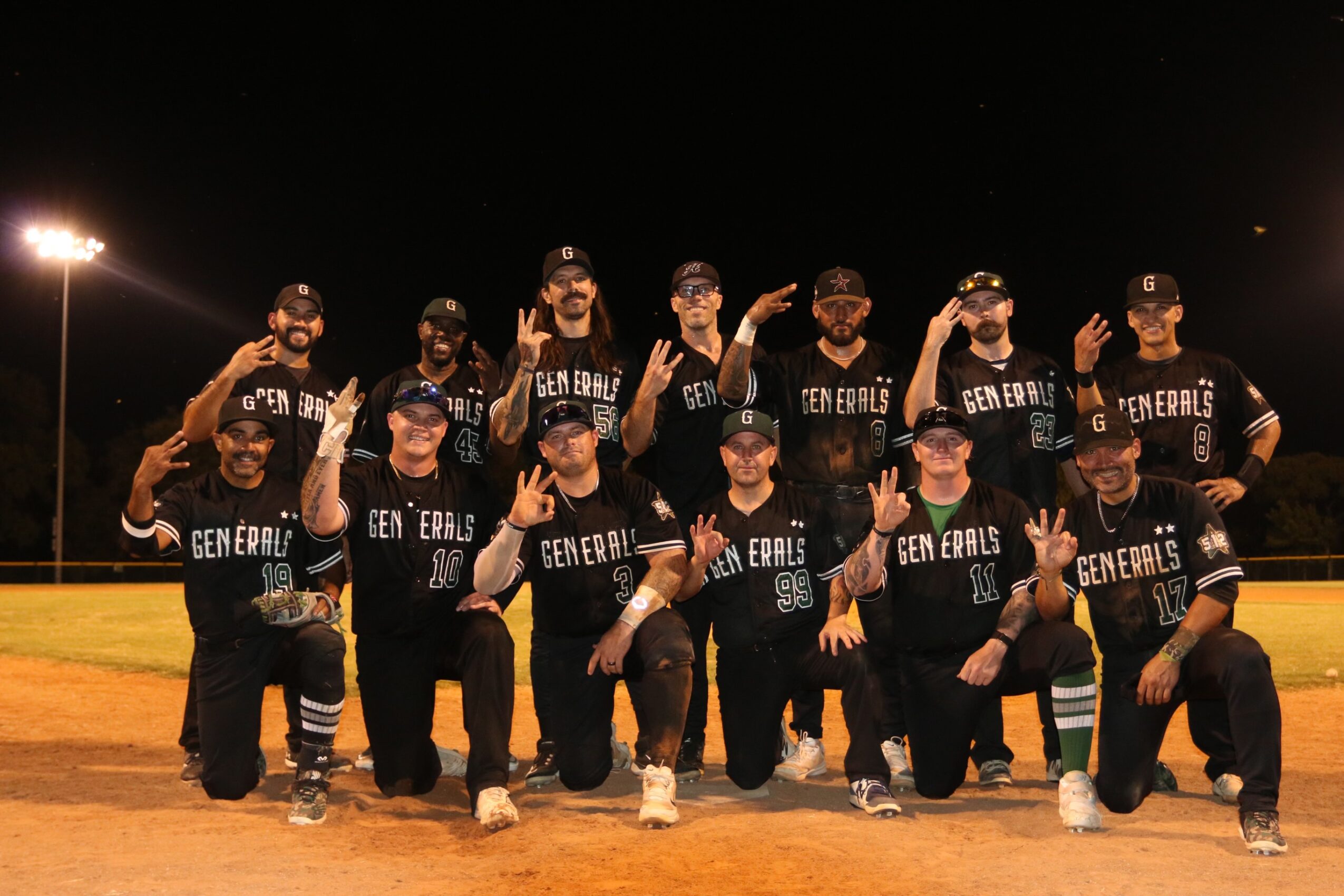 Group of baseball players in uniform posing on field at night, some making peace signs, under bright stadium lights.
