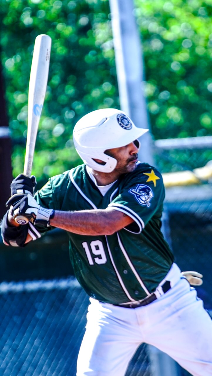 Baseball player in uniform holding bat, wearing helmet, ready to swing, outdoors with green background.