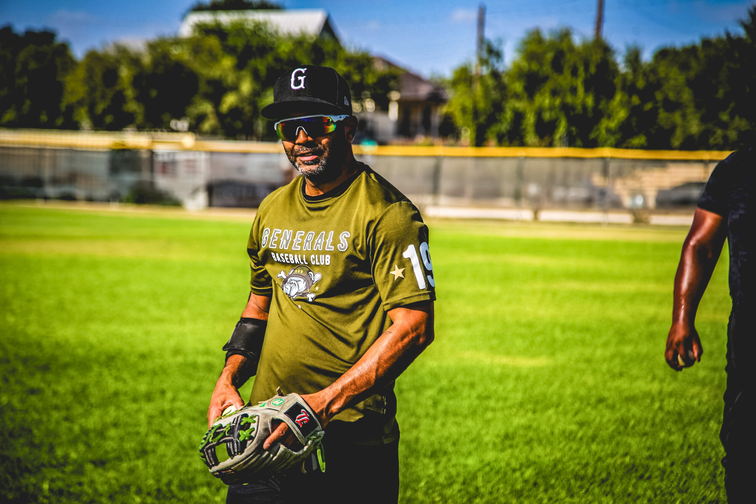 Person wearing a black cap, sunglasses, and a green sports jersey holding a baseball glove on a grassy field.