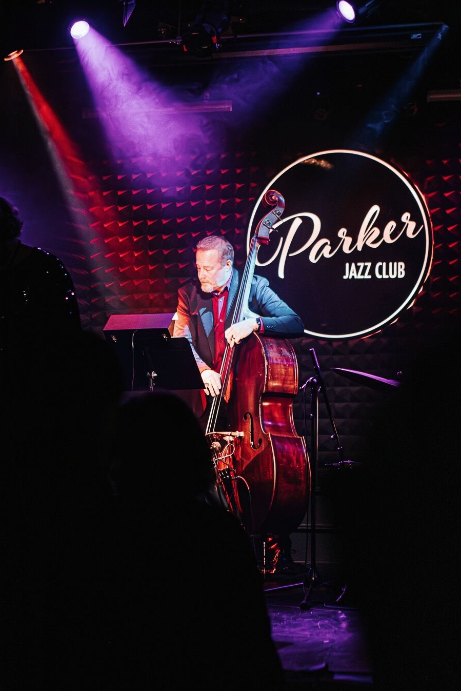 Musician playing double bass on stage with Parker Jazz Club sign in background, purple and red stage lights, dark audience area.