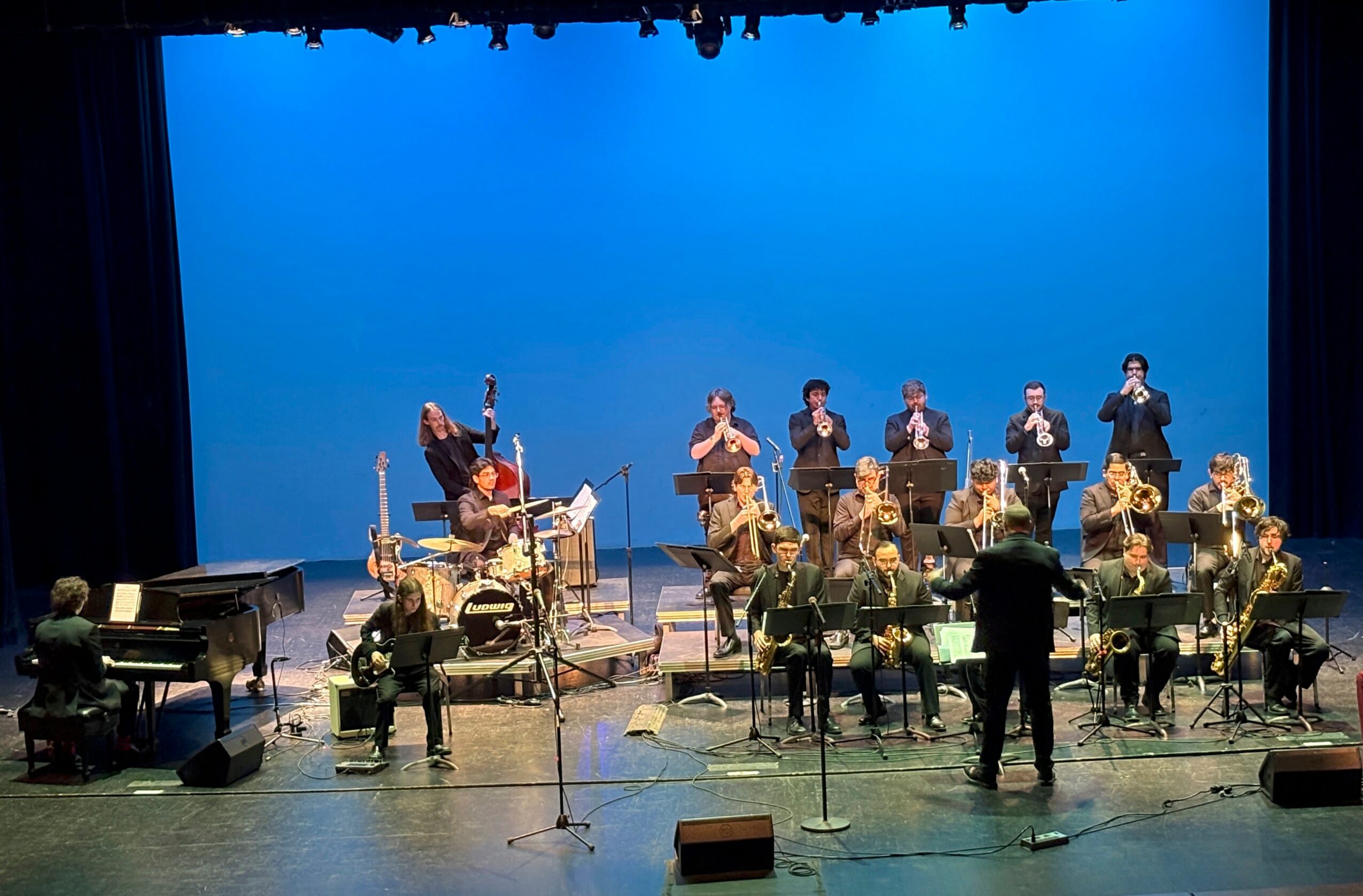 Orchestra performing on stage with musicians playing various instruments, conductor leading, blue backdrop, stage lighting.