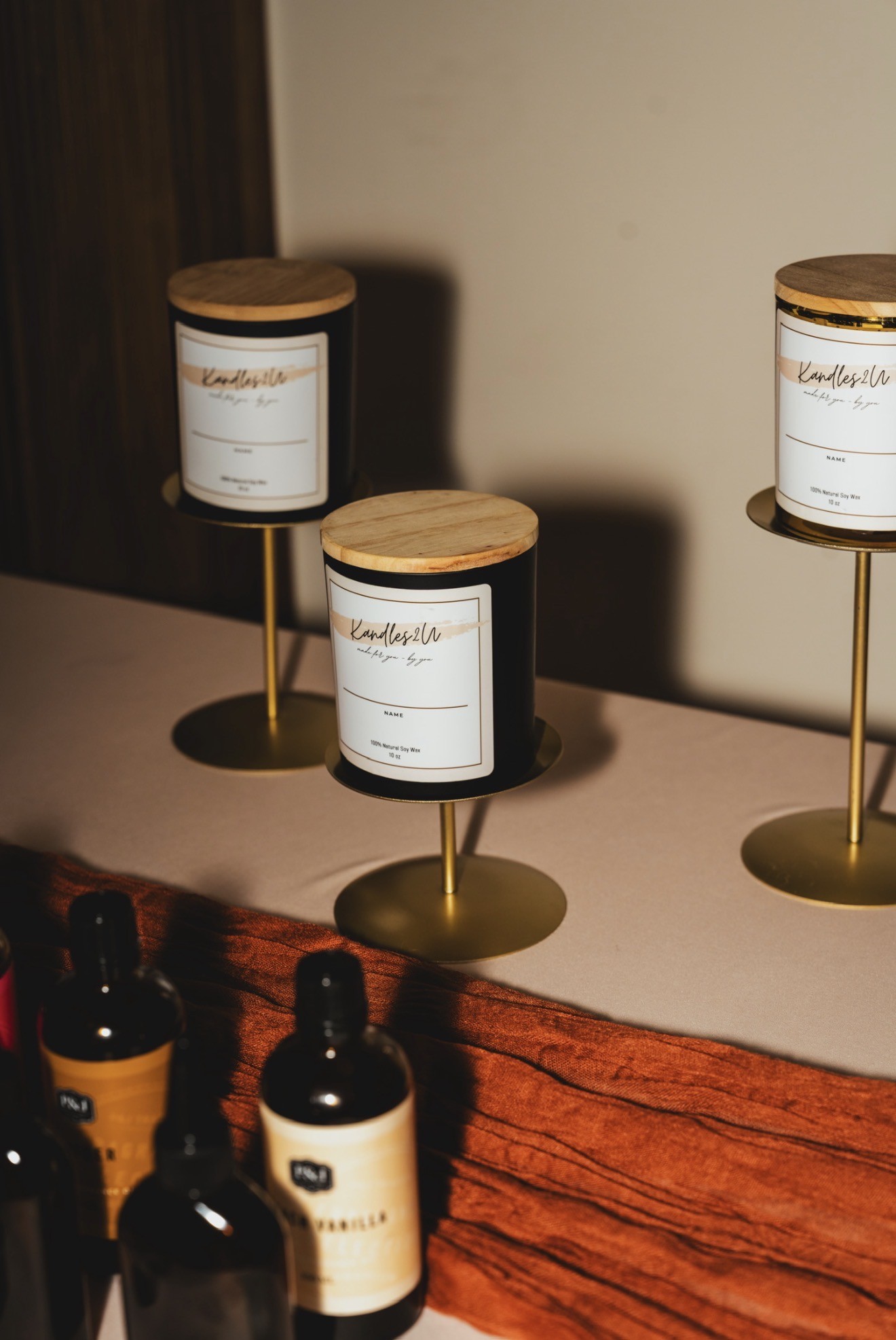 Three candles on gold stands with wooden lids, displayed on a surface with bottles in the foreground.