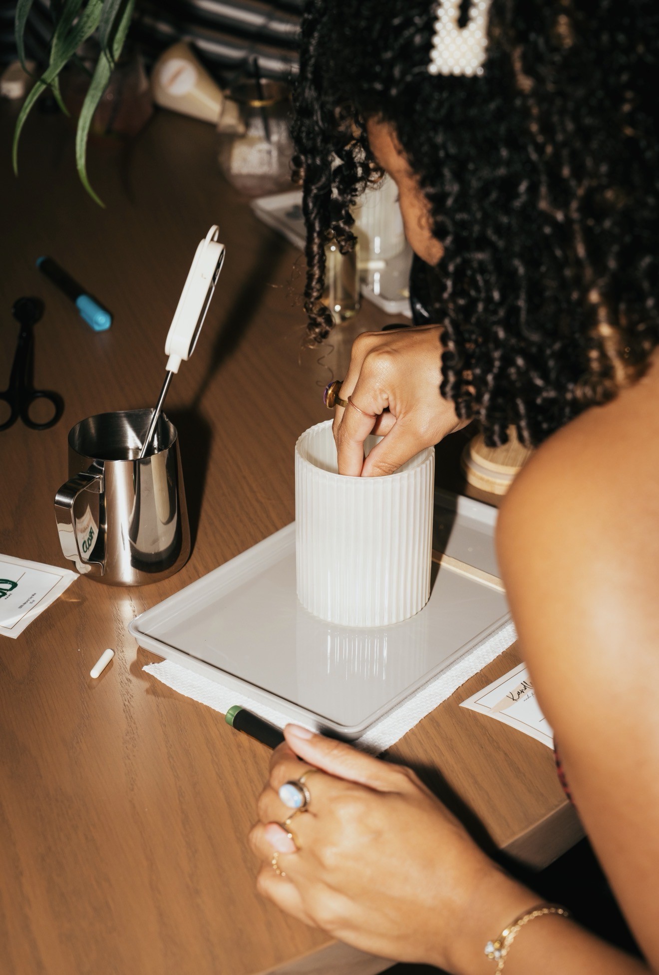 Person with curly hair writing in a white cup on a desk with various office supplies.