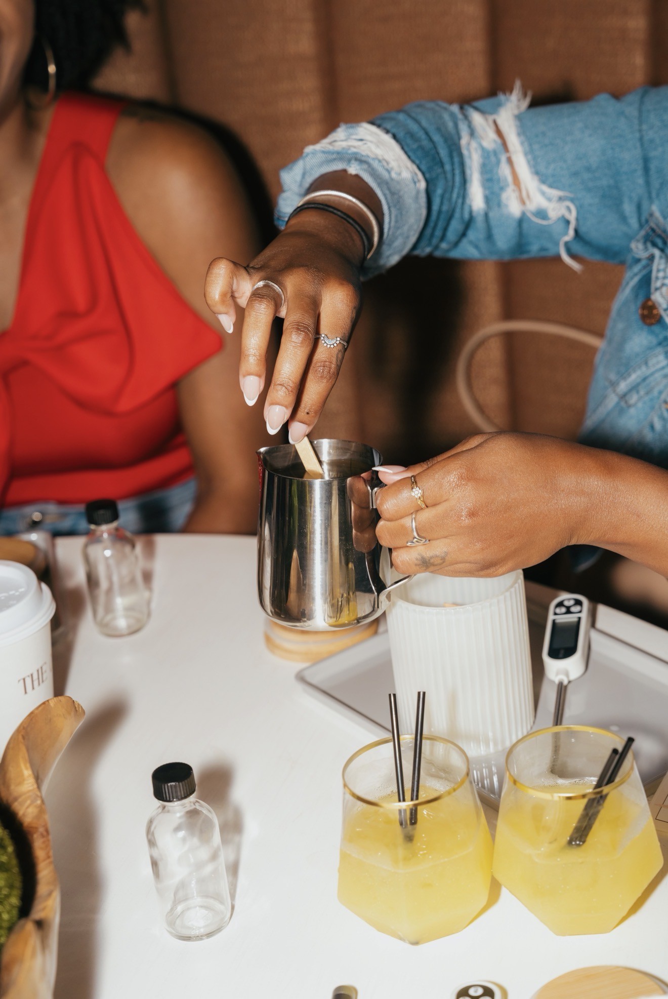 Two people preparing drinks at a table with various bottles and glasses, one person pouring a beverage into a mug.
