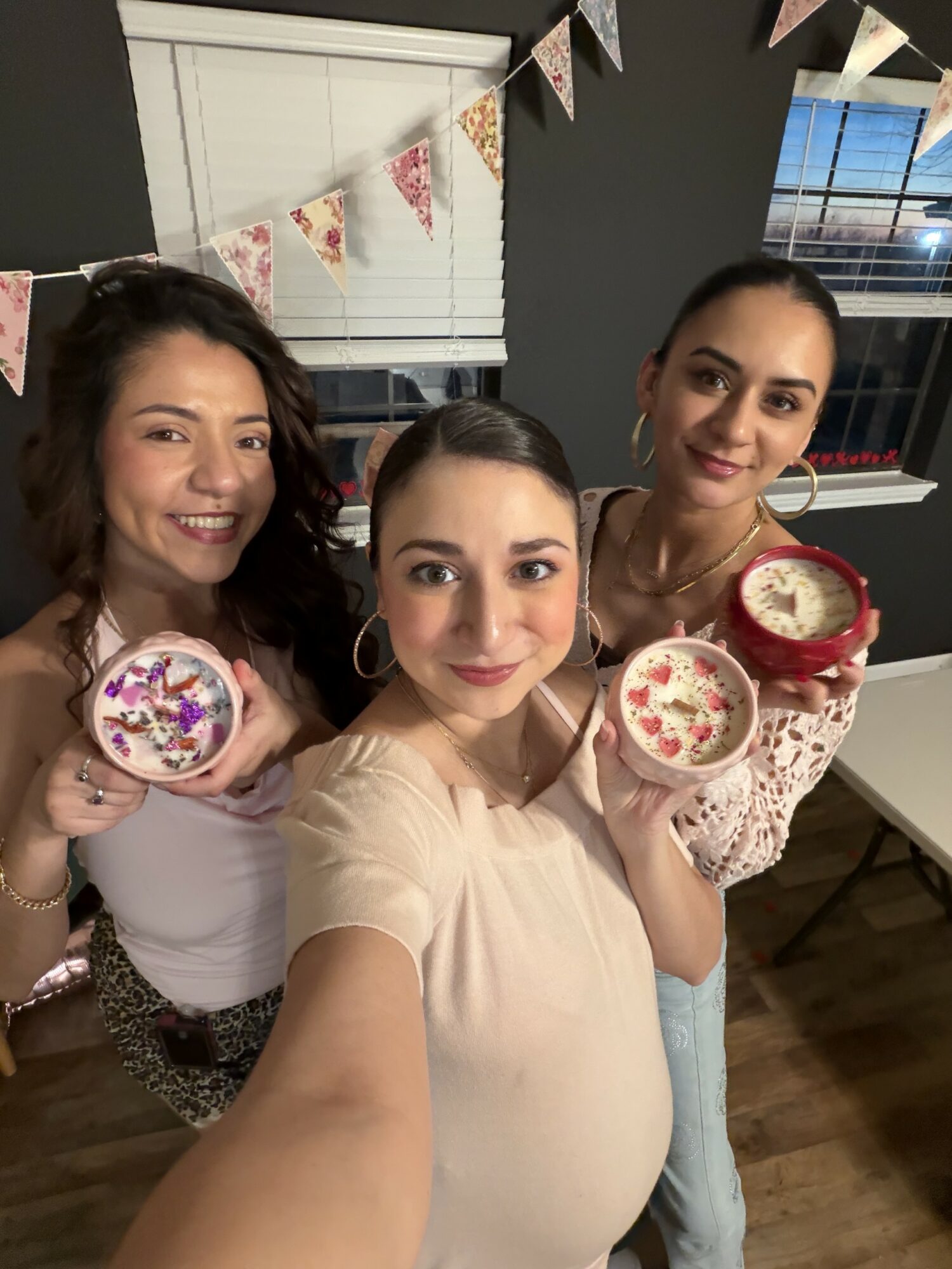Three women smiling indoors, holding decorated bowls, with bunting hanging and a window in the background.