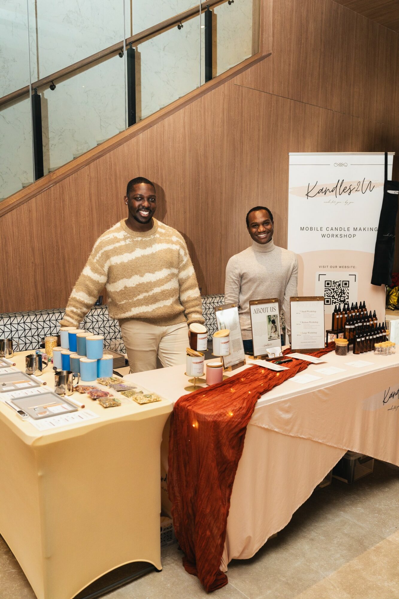 Two smiling men standing behind a table with skincare products and promotional materials inside a wooden-finished room.