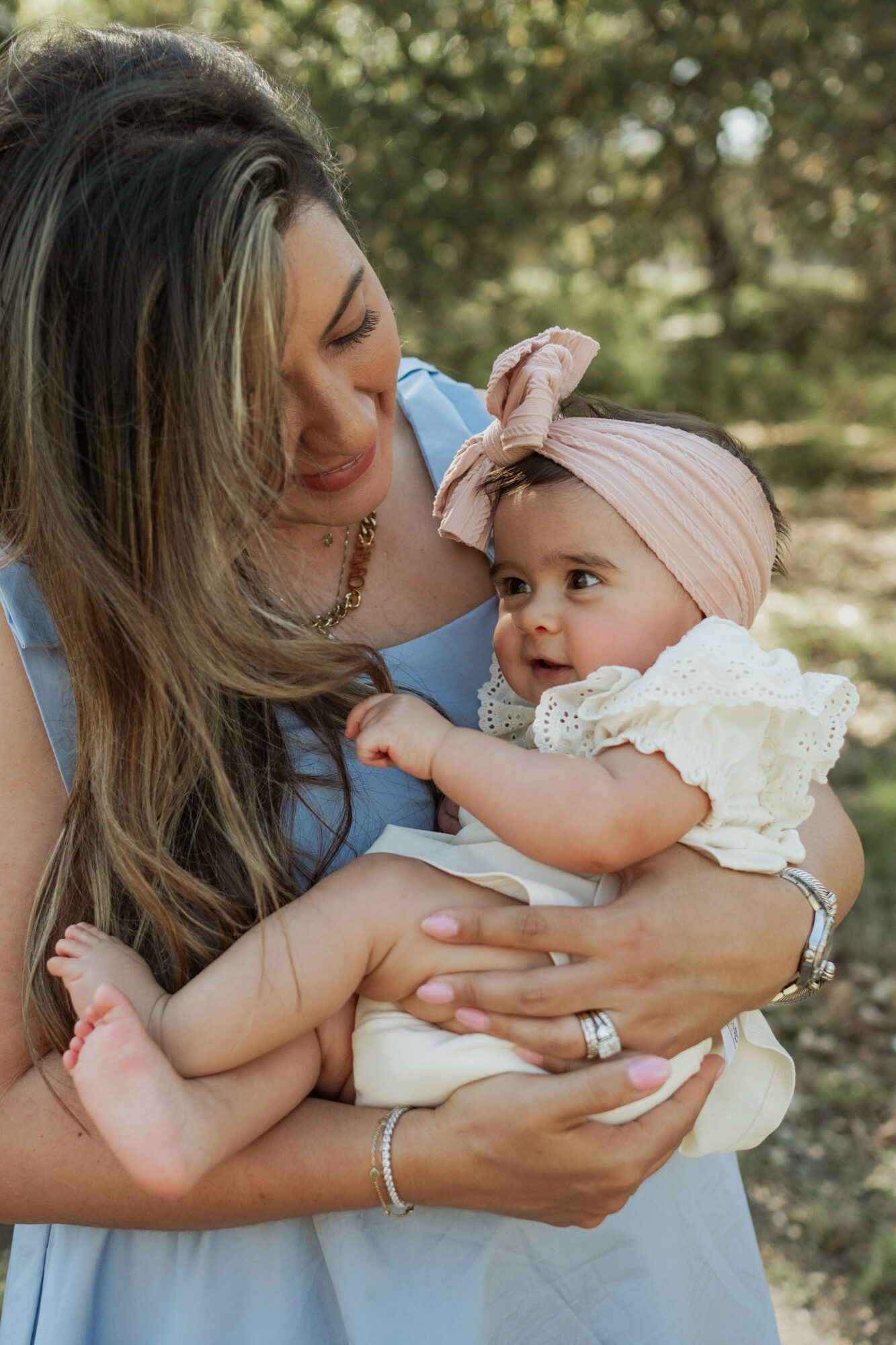 Woman holding a baby girl outdoors with trees in background, woman smiling at the child, who wears a headband and white dress.