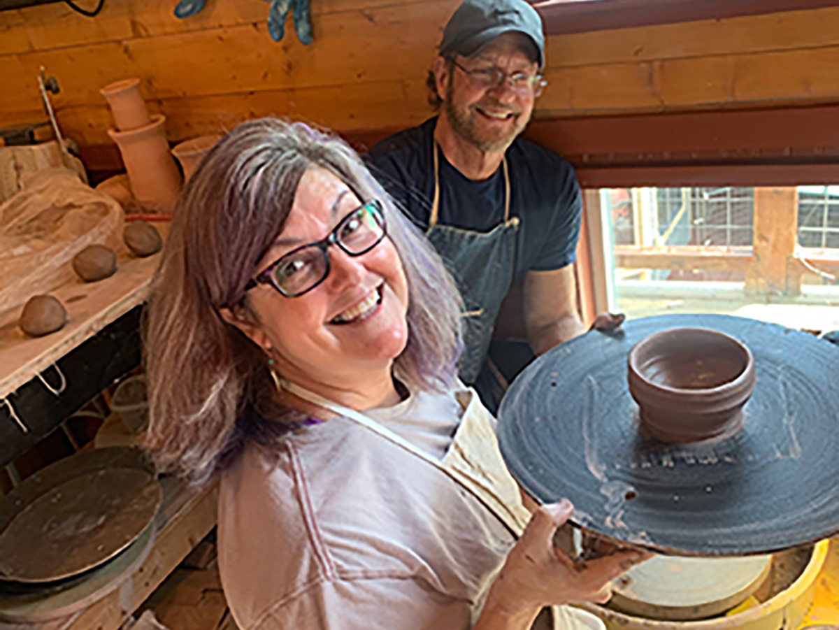 Two people smiling, one holding a pottery wheel with a bowl, in a wooden workshop.