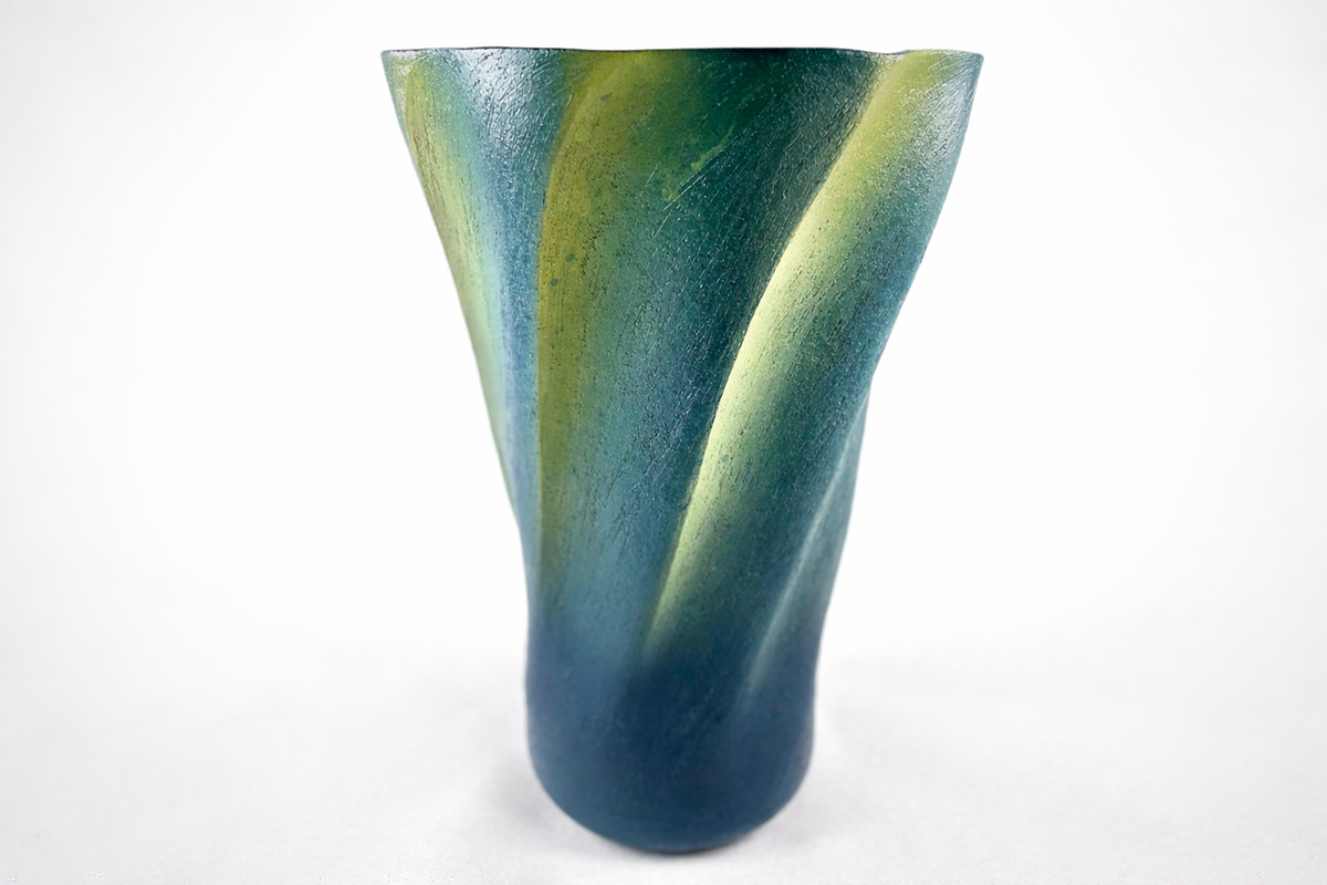 Colorful ceramic vase with green, yellow, and blue hues, tapering towards the bottom, on a white background.