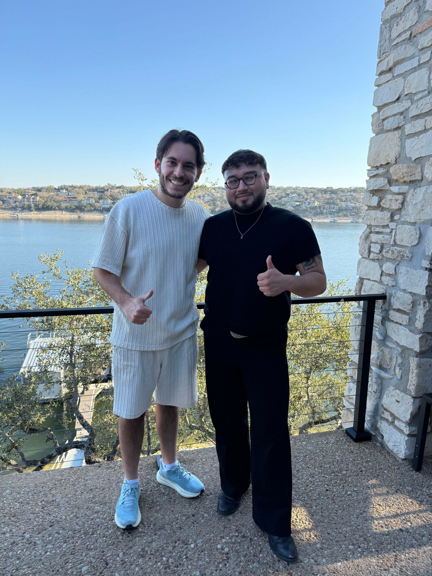 Two men standing outdoors near a railing with a body of water and landscape in the background.