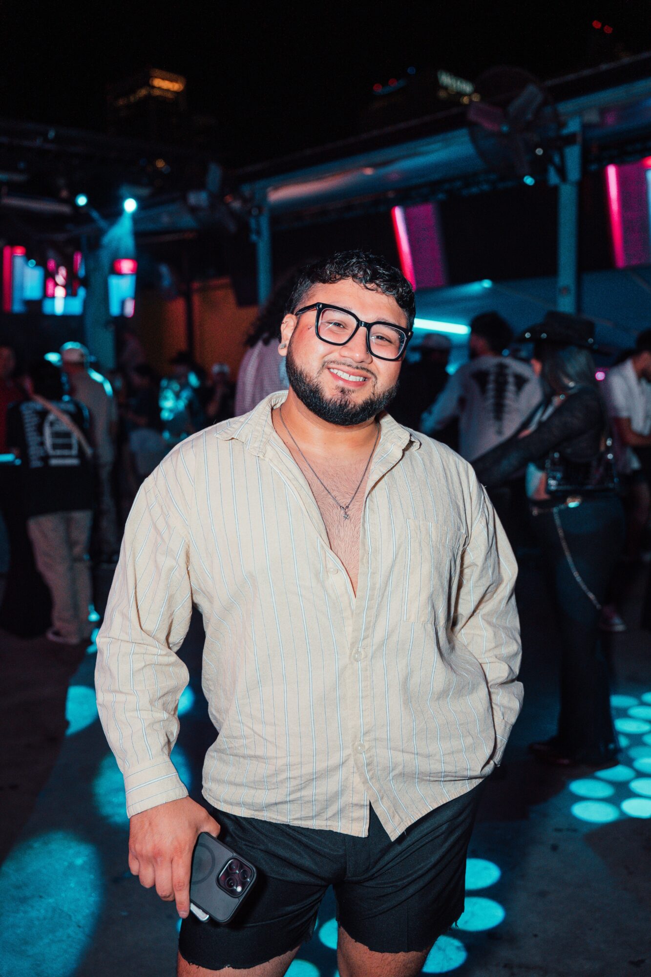 Young man with glasses, beard, smiling, wearing a light-colored shirt and dark shorts, holding a phone, in a nightclub.