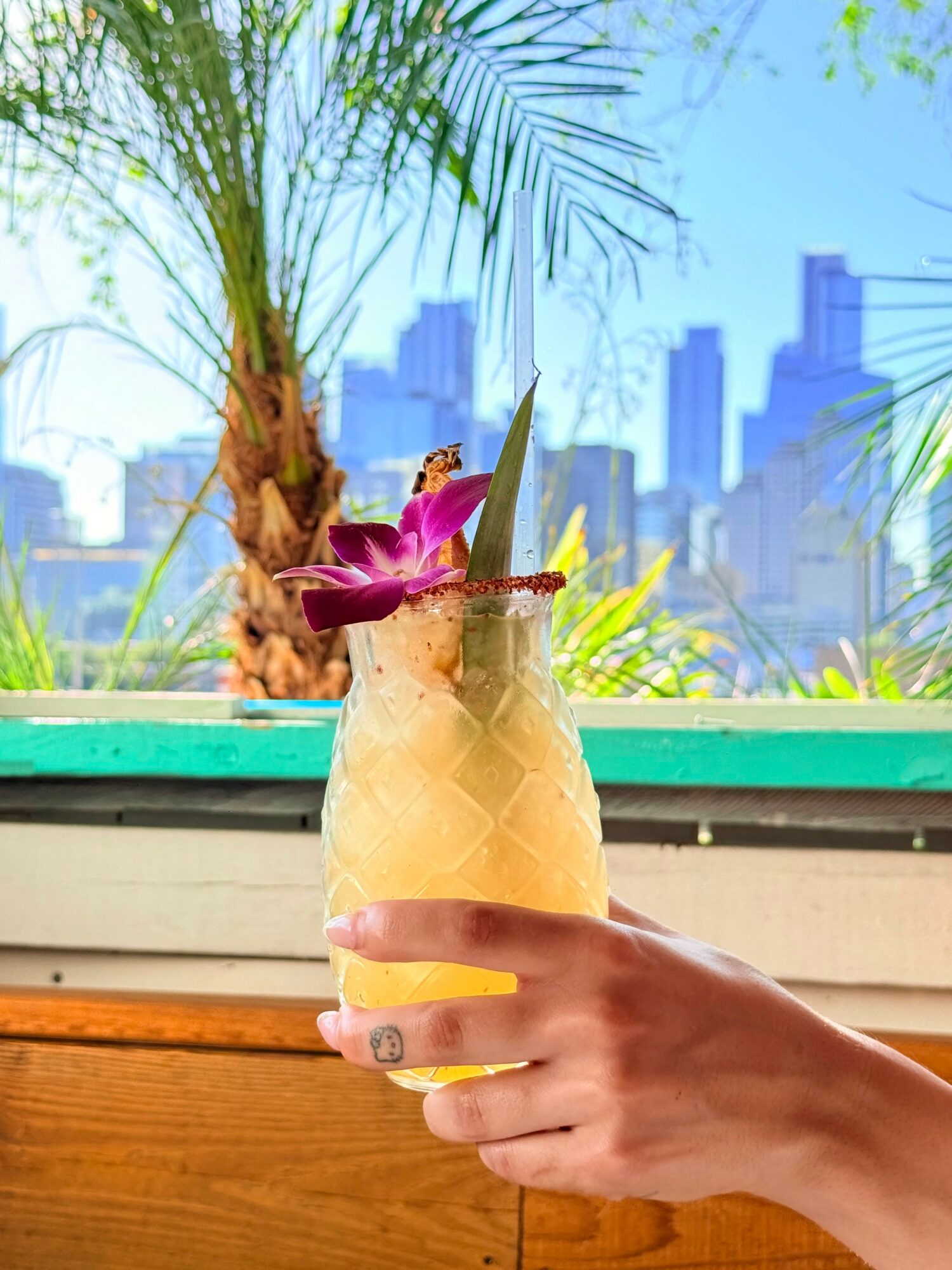 Hand holding a pineapple drink with a straw and flower, city skyline in background, palm tree nearby.