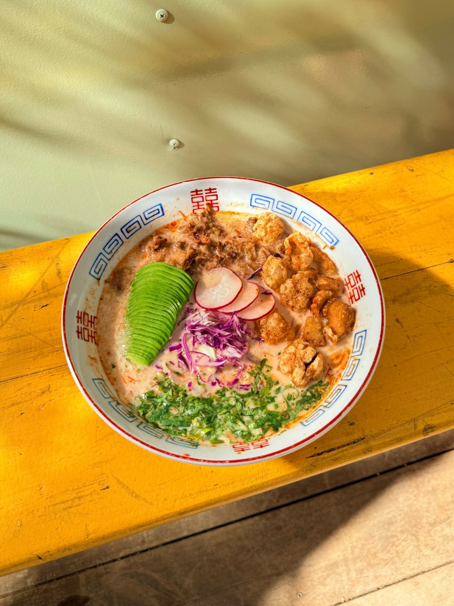Bowl of noodle soup with sliced avocado, radish, purple cabbage, and green herbs on a wooden surface.