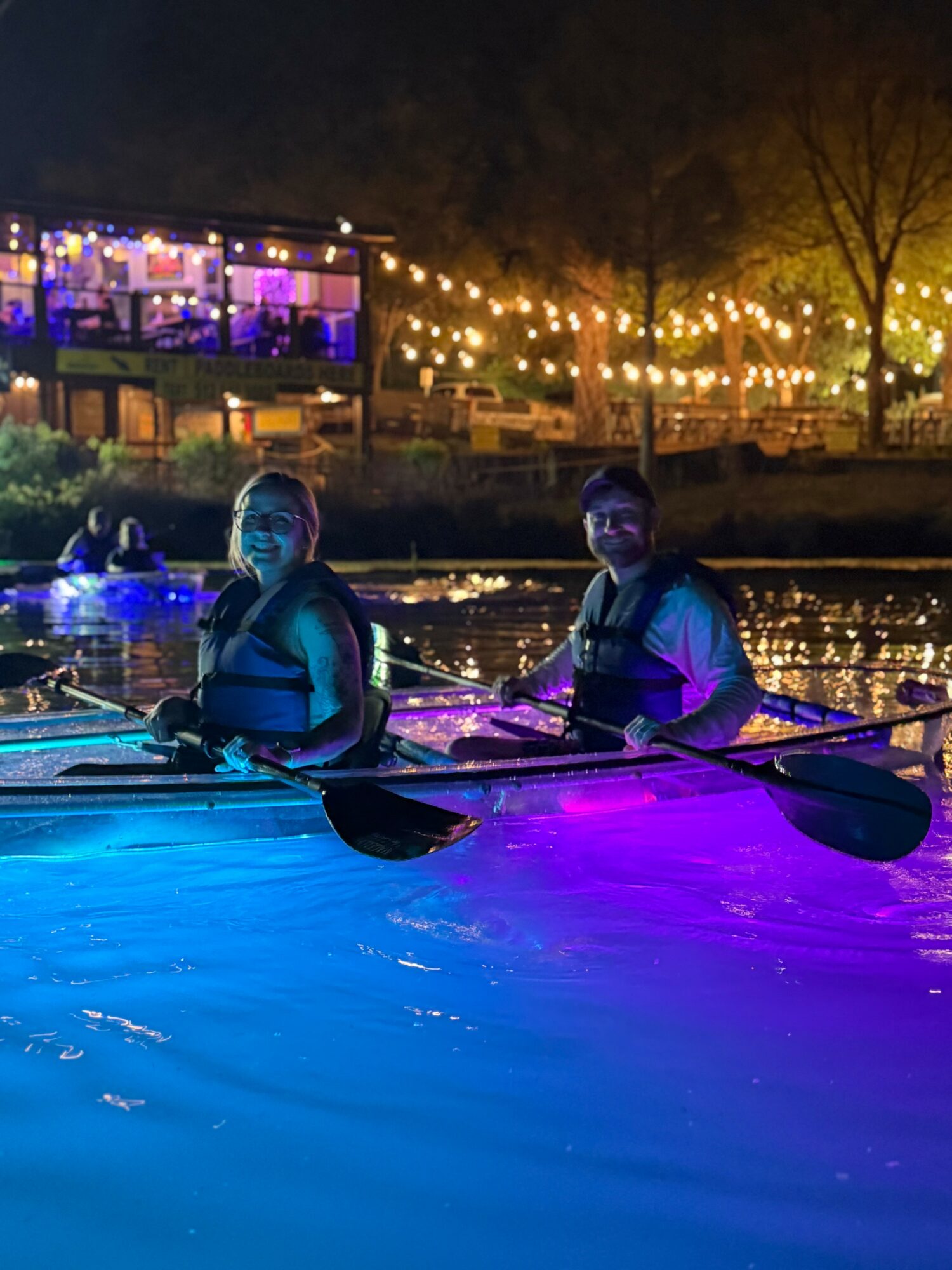 People in kayaks on water at night with colorful lights and trees in background.