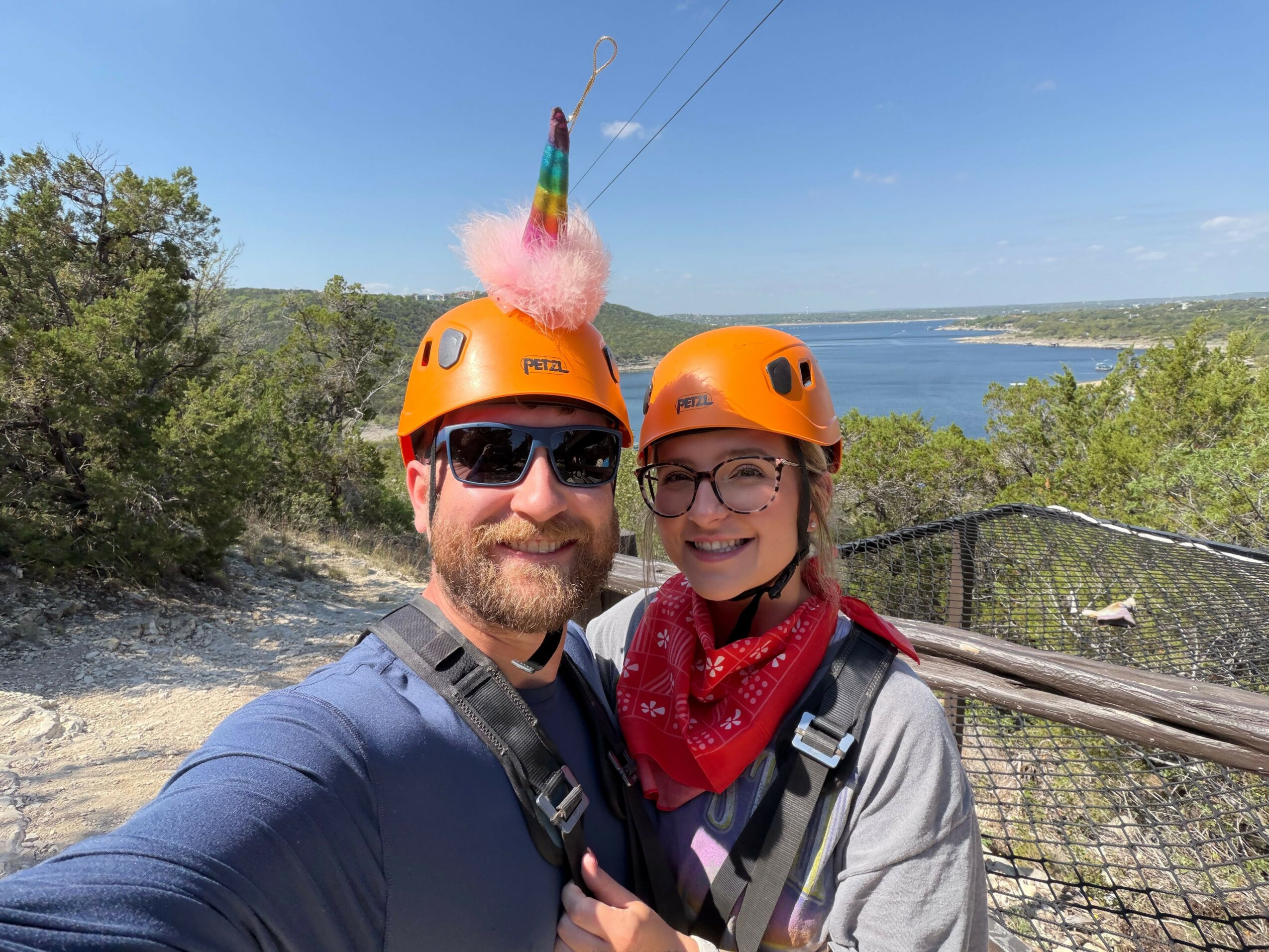 Two people wearing orange helmets and sunglasses taking a selfie outdoors with a lake and trees in the background.