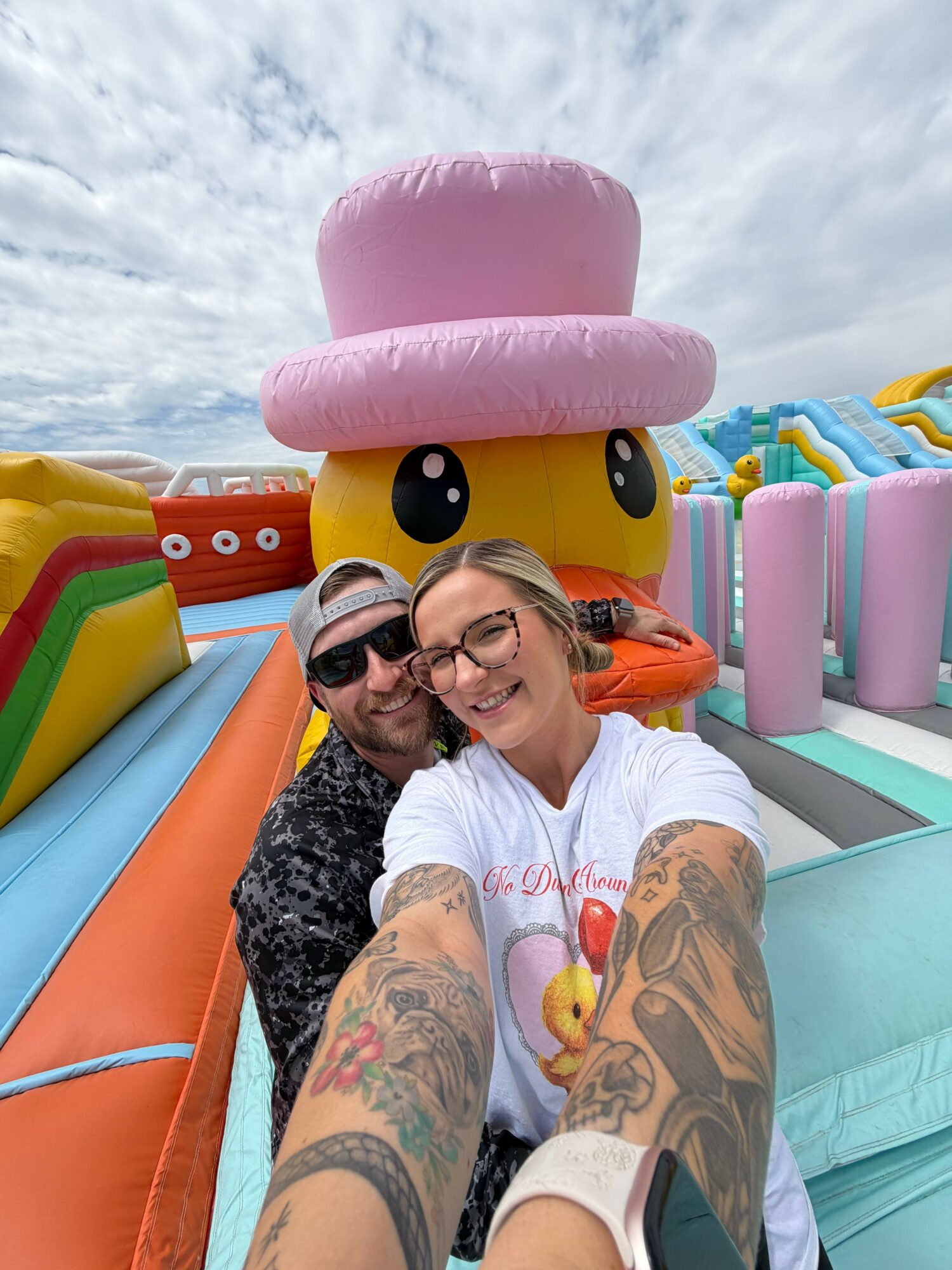 Two people taking a selfie in front of a colorful inflatable slide with a large pink hat-shaped decoration, outdoors on a cloudy day.