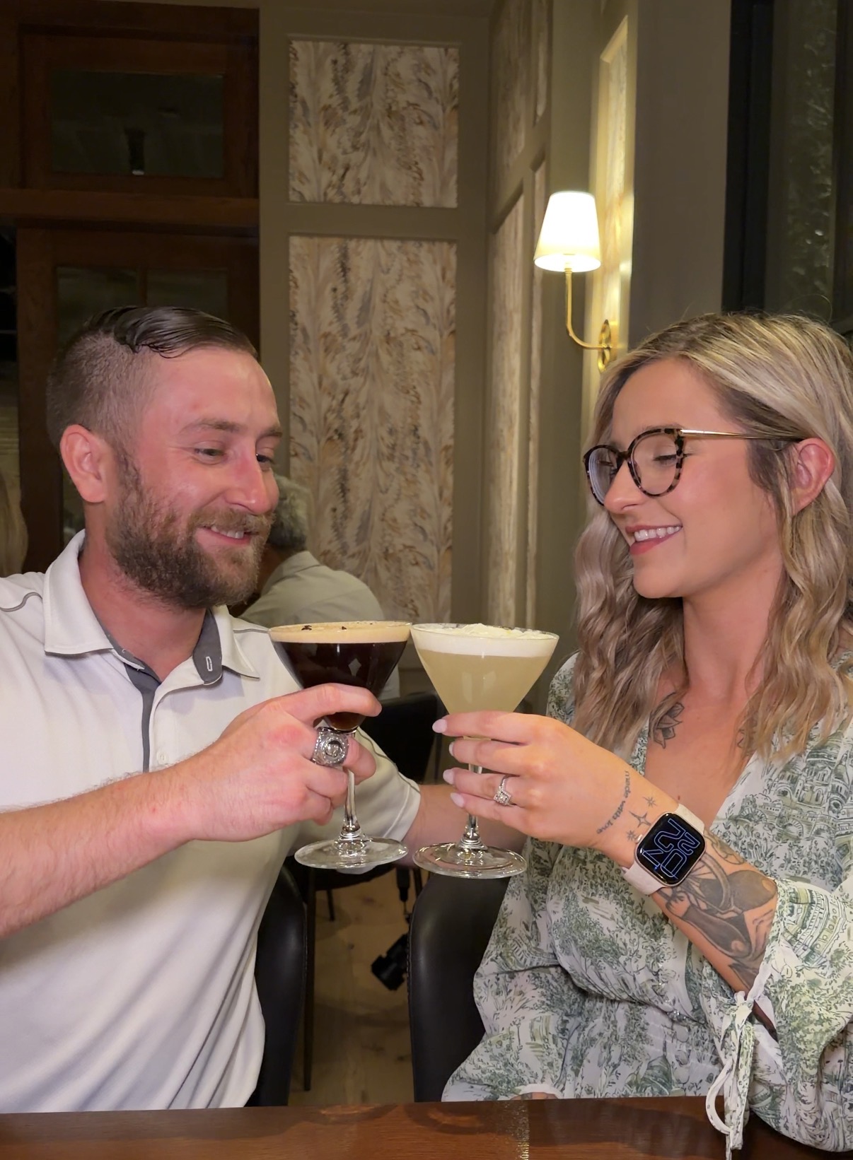 A man and woman toast with cocktails in a cozy indoor setting, smiling at each other.