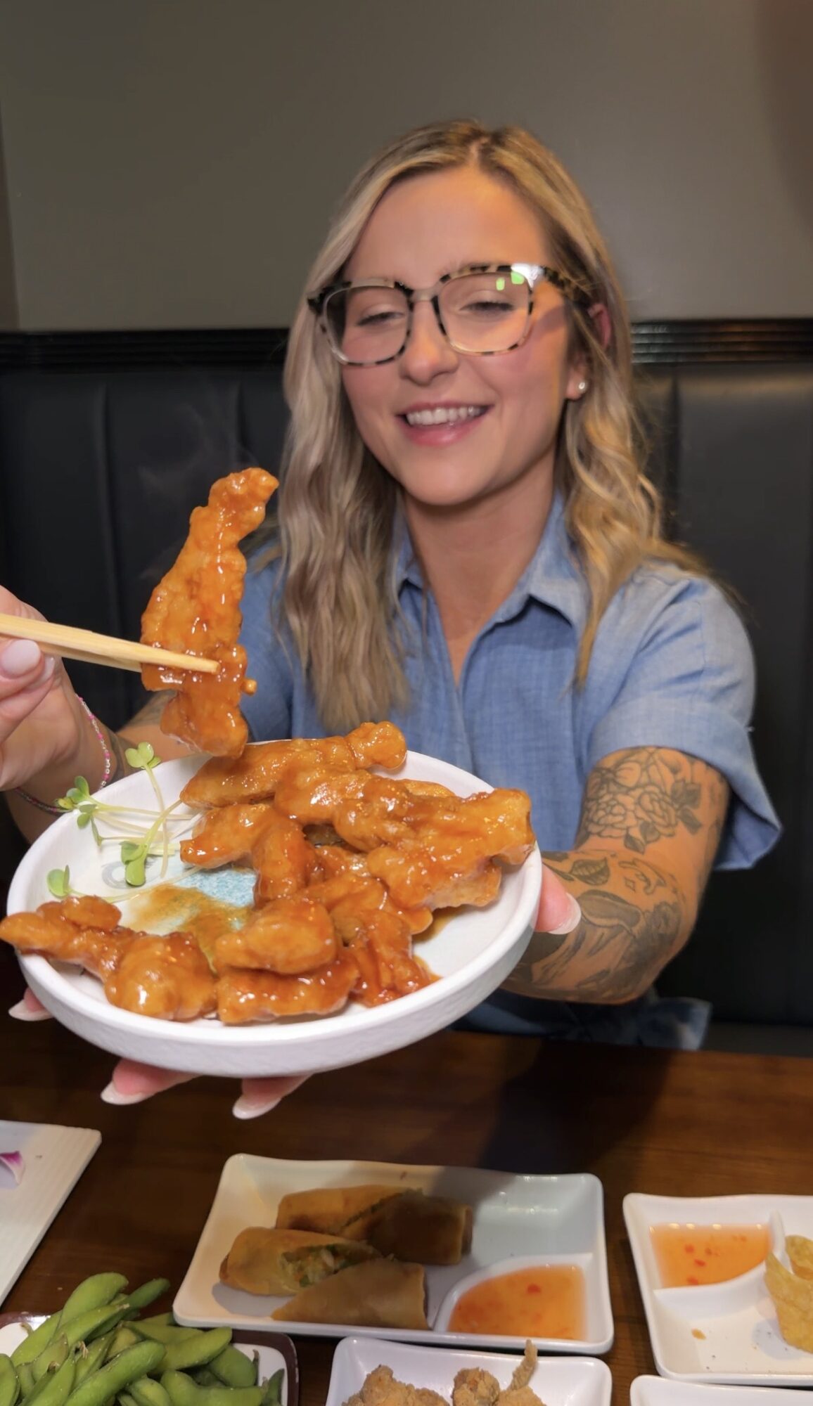 Young woman with glasses and blonde hair holding a plate of orange chicken at a restaurant.