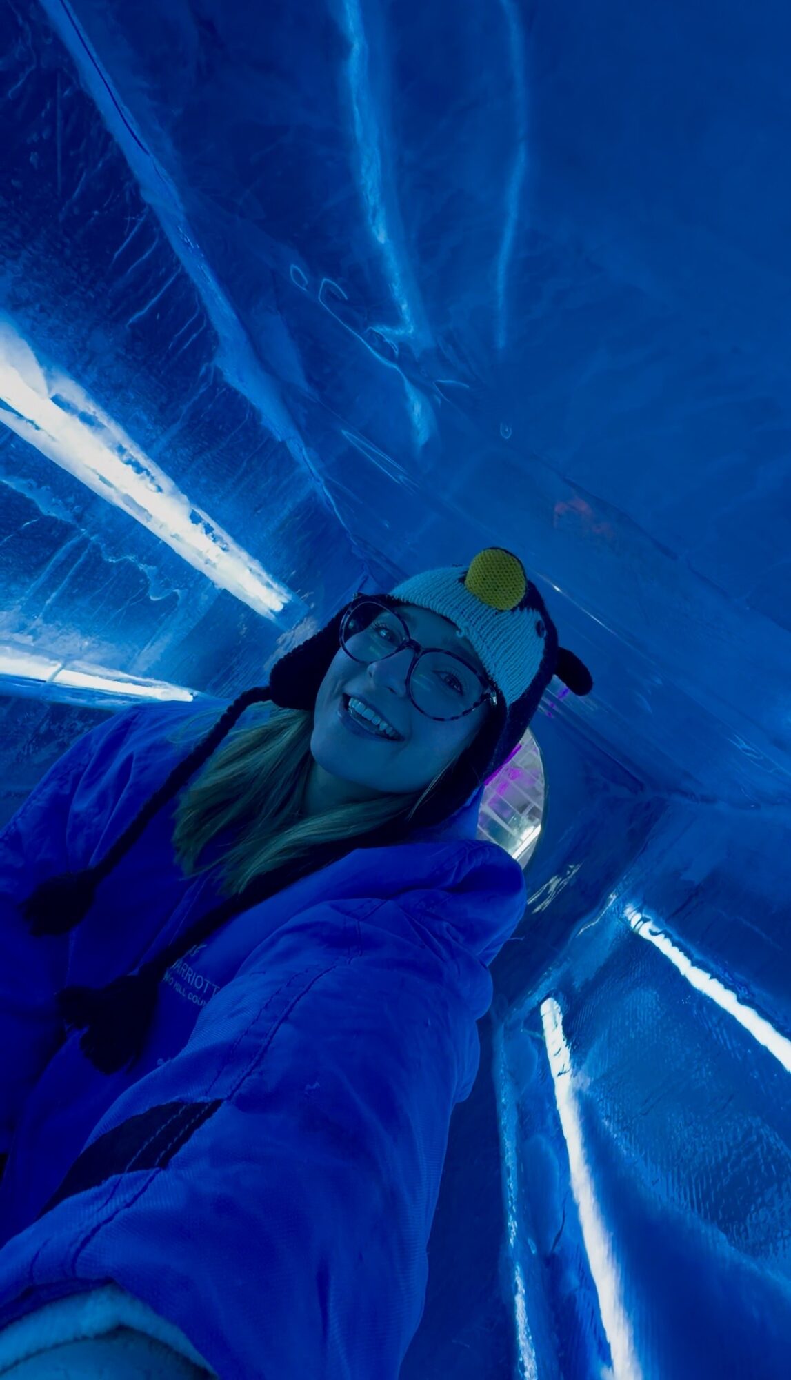 Person wearing safety gear and glasses, smiling, in a tunnel with blue lighting and reflective surfaces.
