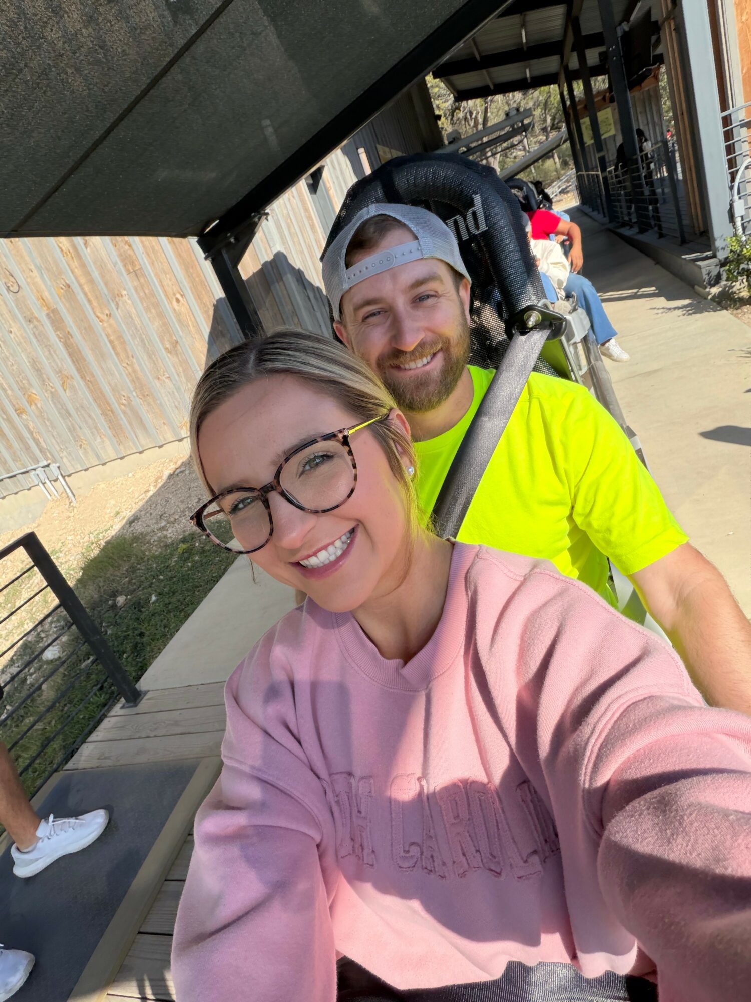 Two smiling people taking a selfie outdoors on a sunny day, with a woman in glasses and a pink sweatshirt and a man in a neon yellow shirt and cap.