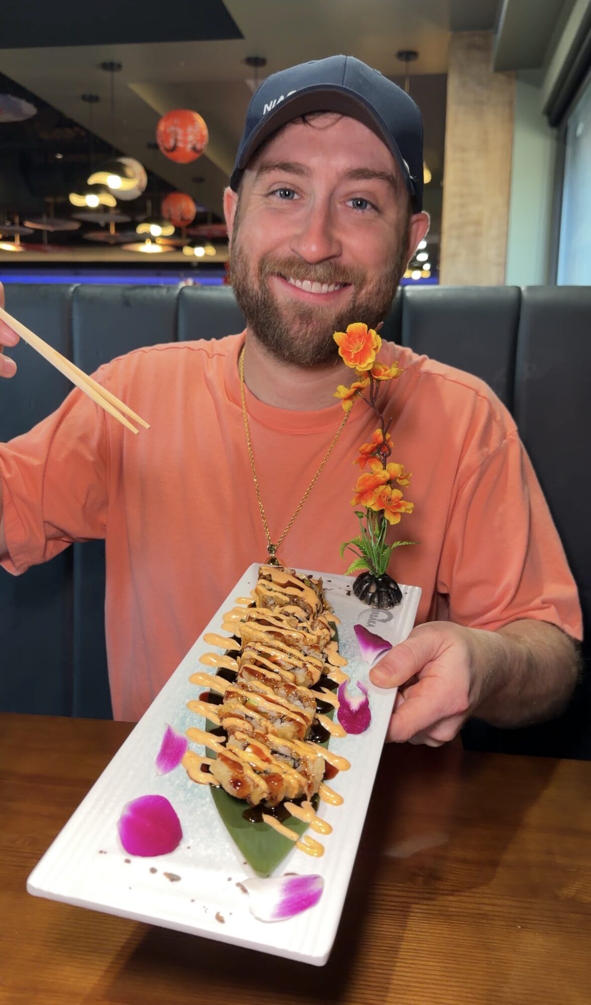 Smiling man with beard and cap holding a plate of sushi with flowers and garnishes.