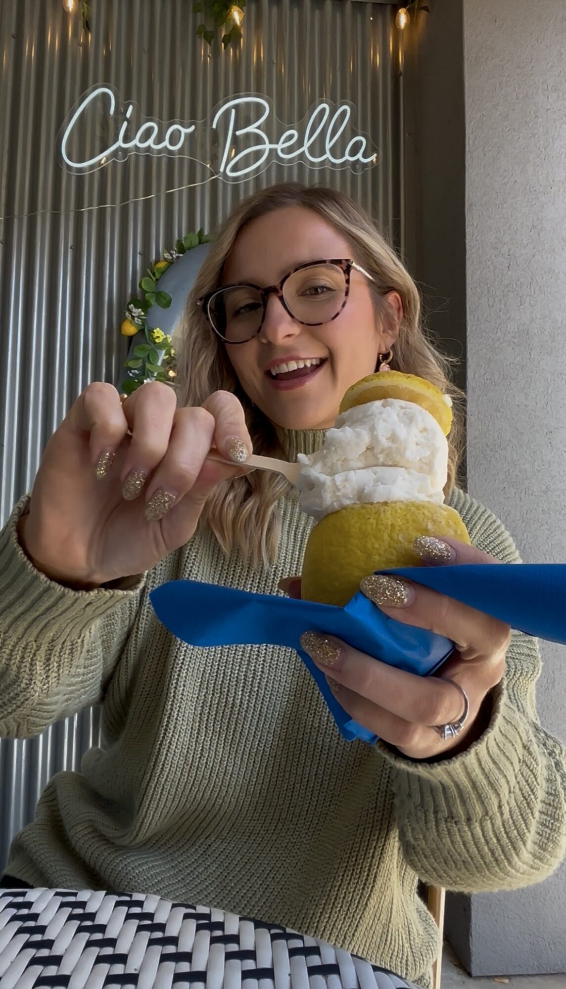 Young woman with glasses smiling, holding a spoon with ice cream, in front of a sign that says 'Ciao Bella'.