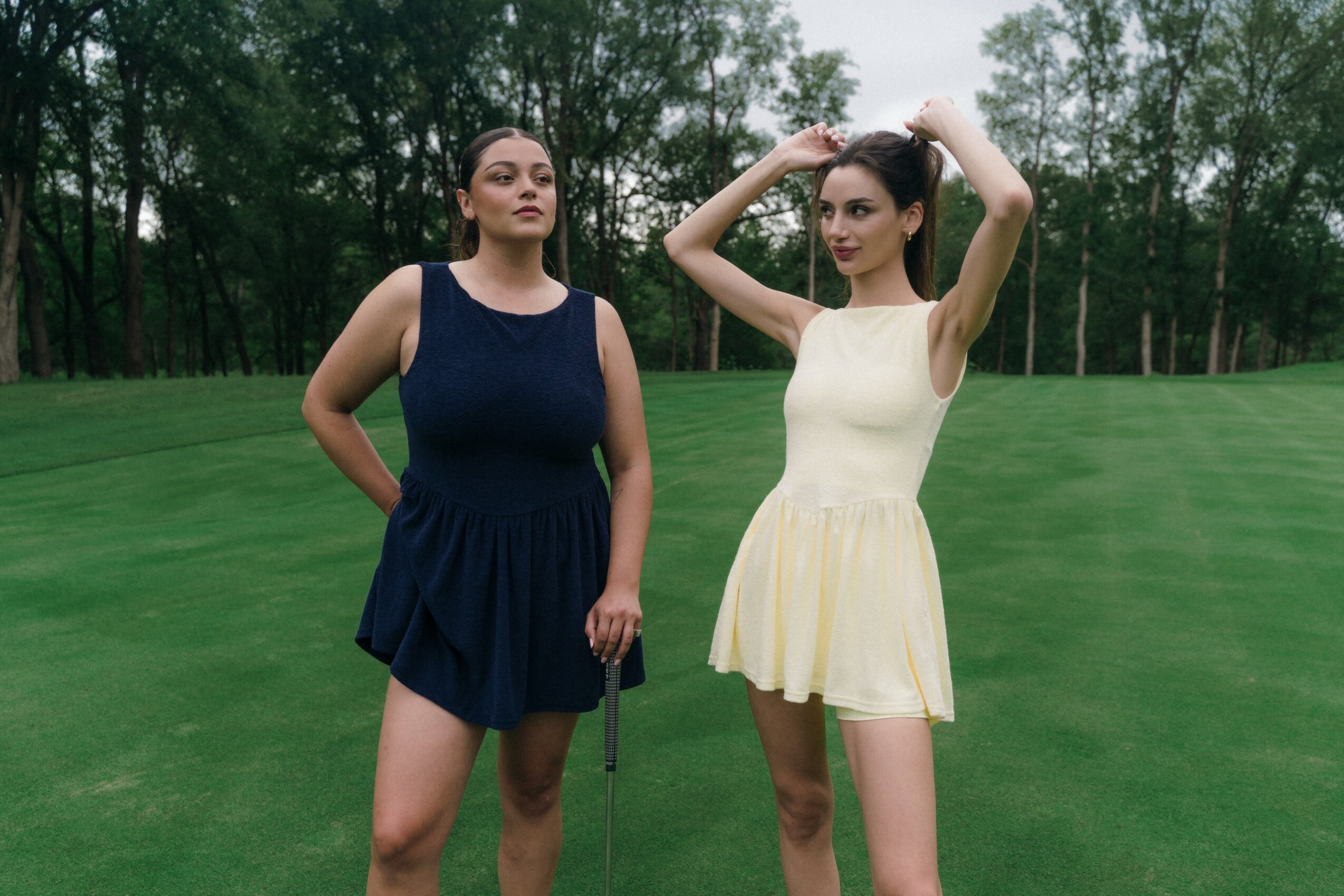 Two women standing outdoors on grass with trees in background, one in navy dress, other in light yellow dress, posing.