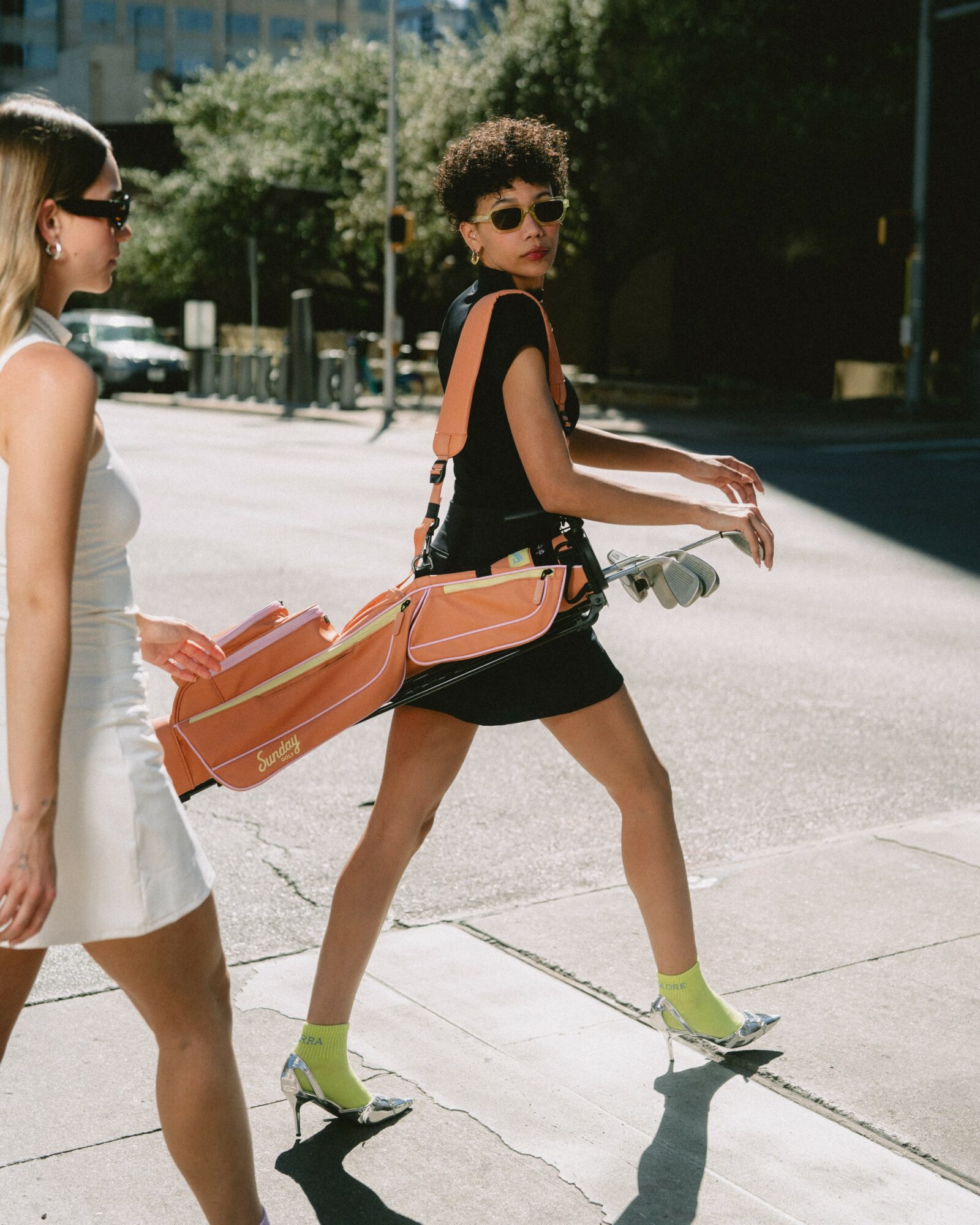 Young woman with sunglasses and a black dress walking with a skateboard on a city sidewalk.