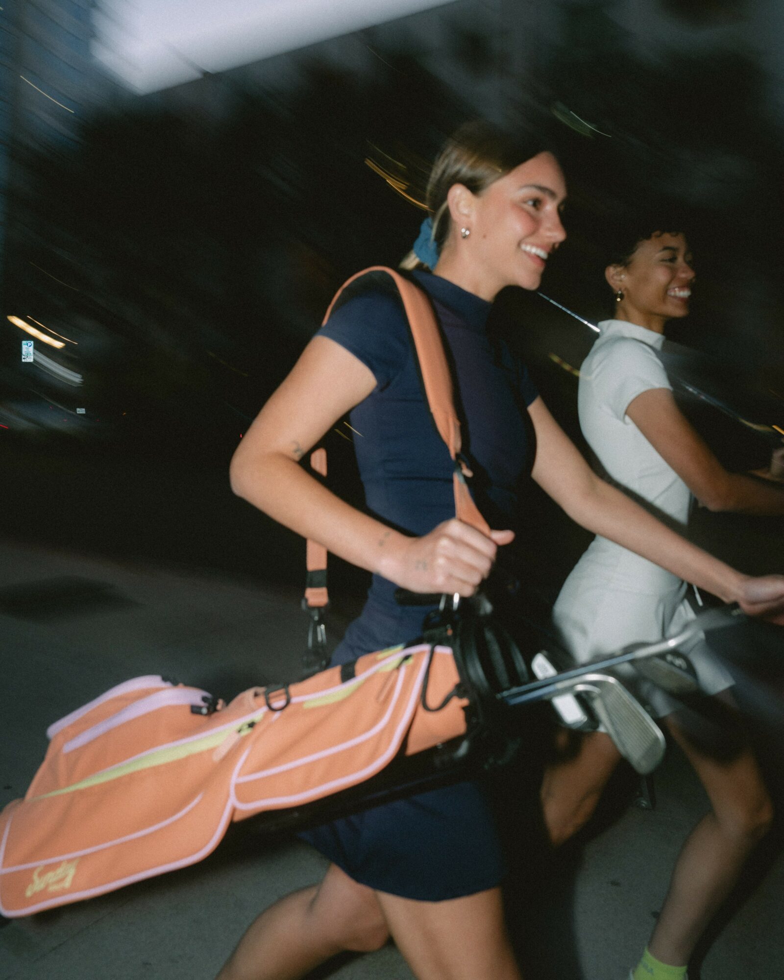 Two women walking outdoors at night, smiling, with one carrying a bag and the other holding a bicycle handlebar.