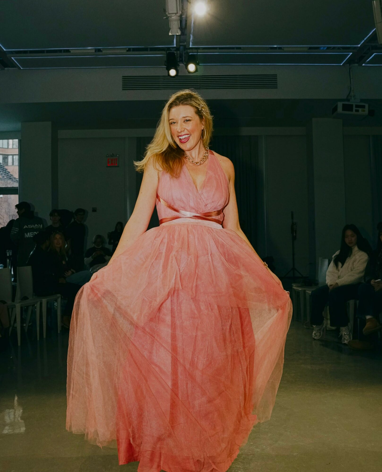 Woman in pink dress smiling, holding skirt, in indoor setting with audience seated around.