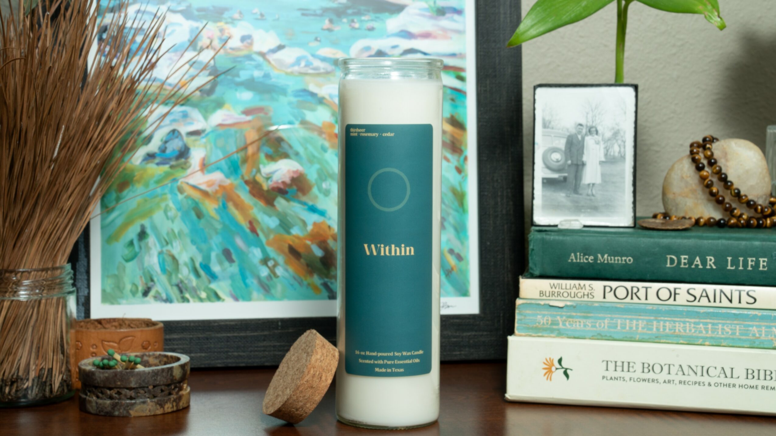 White candle with teal label on a wooden surface, surrounded by books, a framed painting, and decorative items.