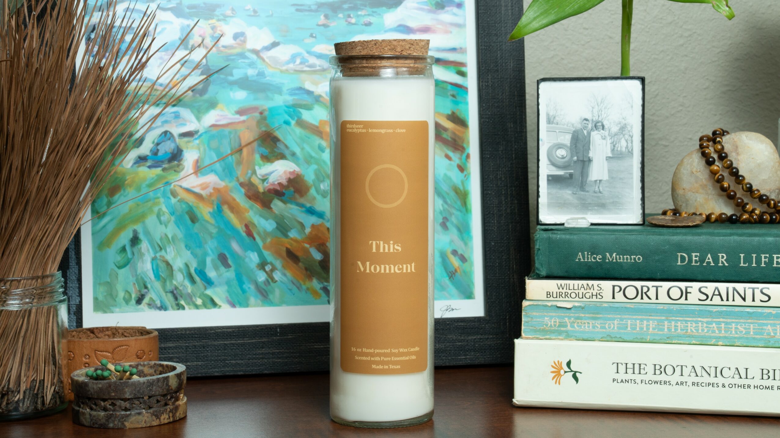 Candle in a glass container with a wooden lid, surrounded by books, a framed painting, and decorative items on a shelf.