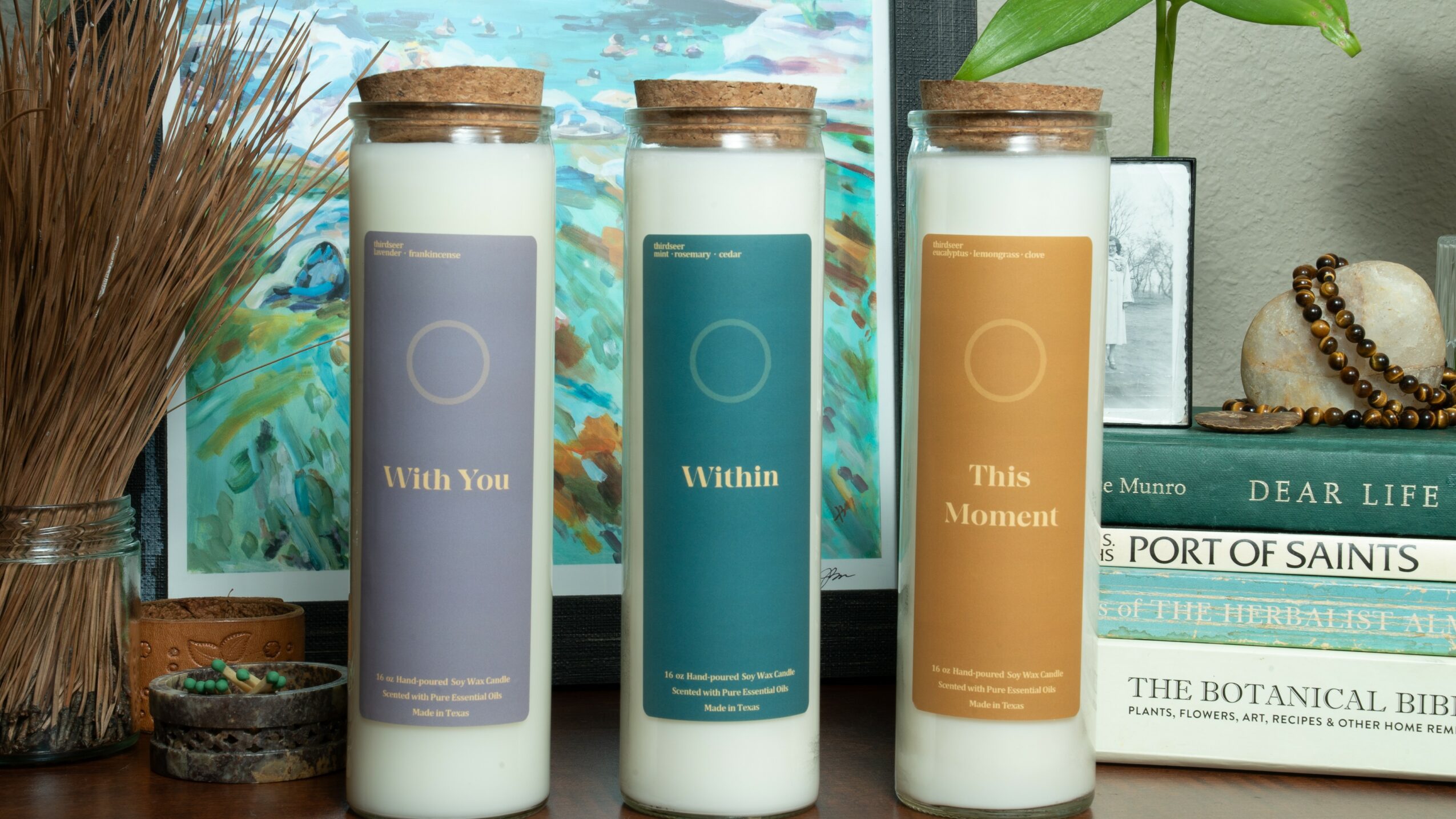 Three candles in glass jars with wooden lids, arranged on a shelf with books and decorative items.
