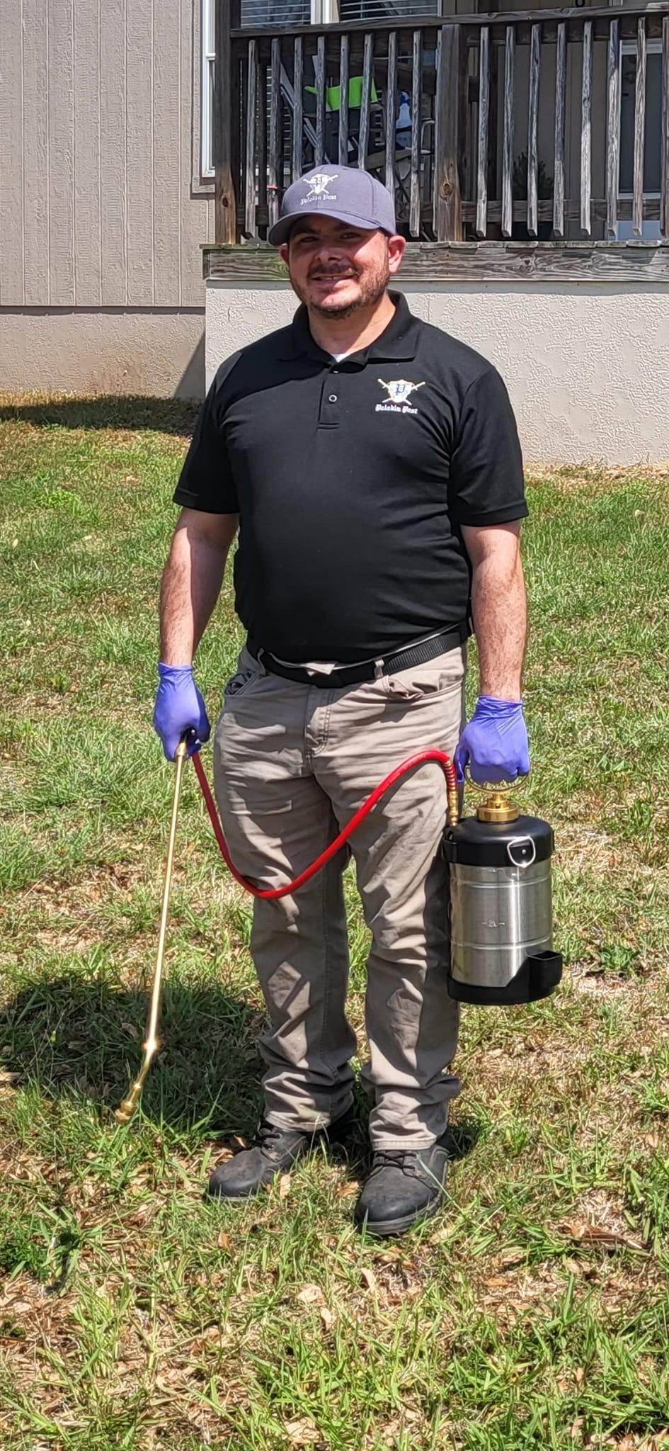 Man standing outdoors holding a device with a hose, wearing gloves, a black polo shirt, and a cap.