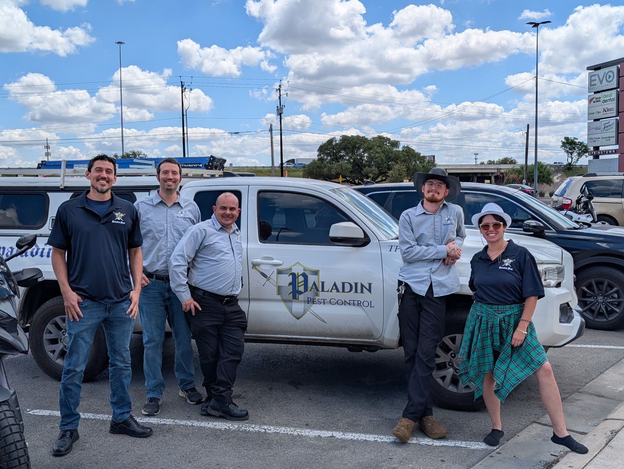 Five people stand in front of a white vehicle with a logo, in a parking lot under a blue sky with clouds.