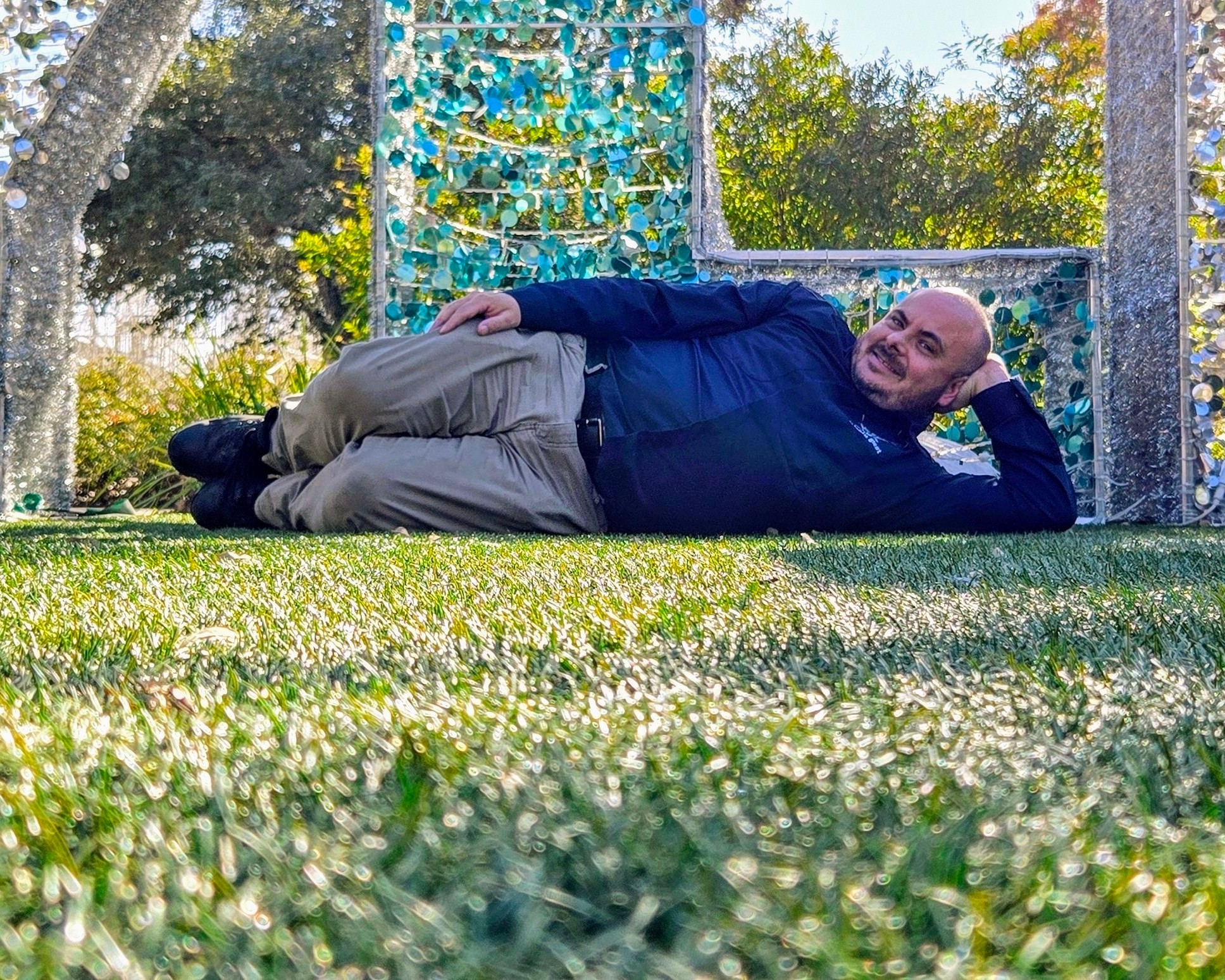 Man lying on grass with colorful decorative structures in background, smiling and resting on his side.