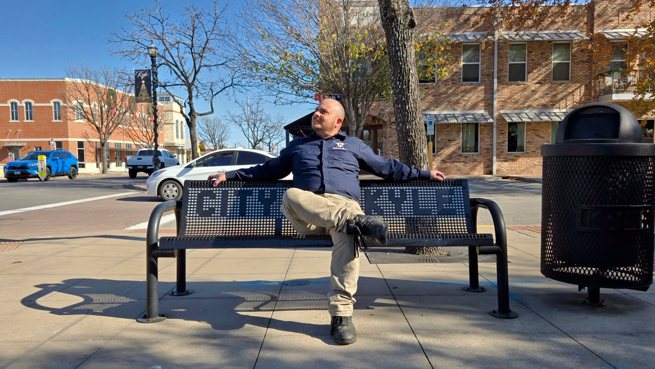 Man sitting on a bench with arms stretched out, in a city park area with trees and buildings in background.
