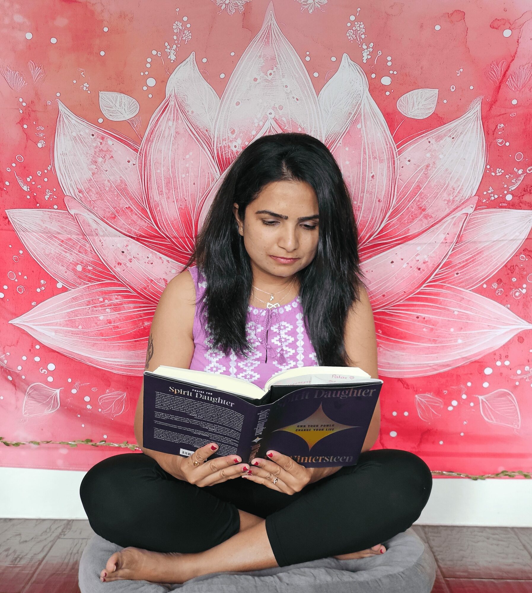 Woman sitting cross-legged reading a book in front of a pink lotus flower backdrop.
